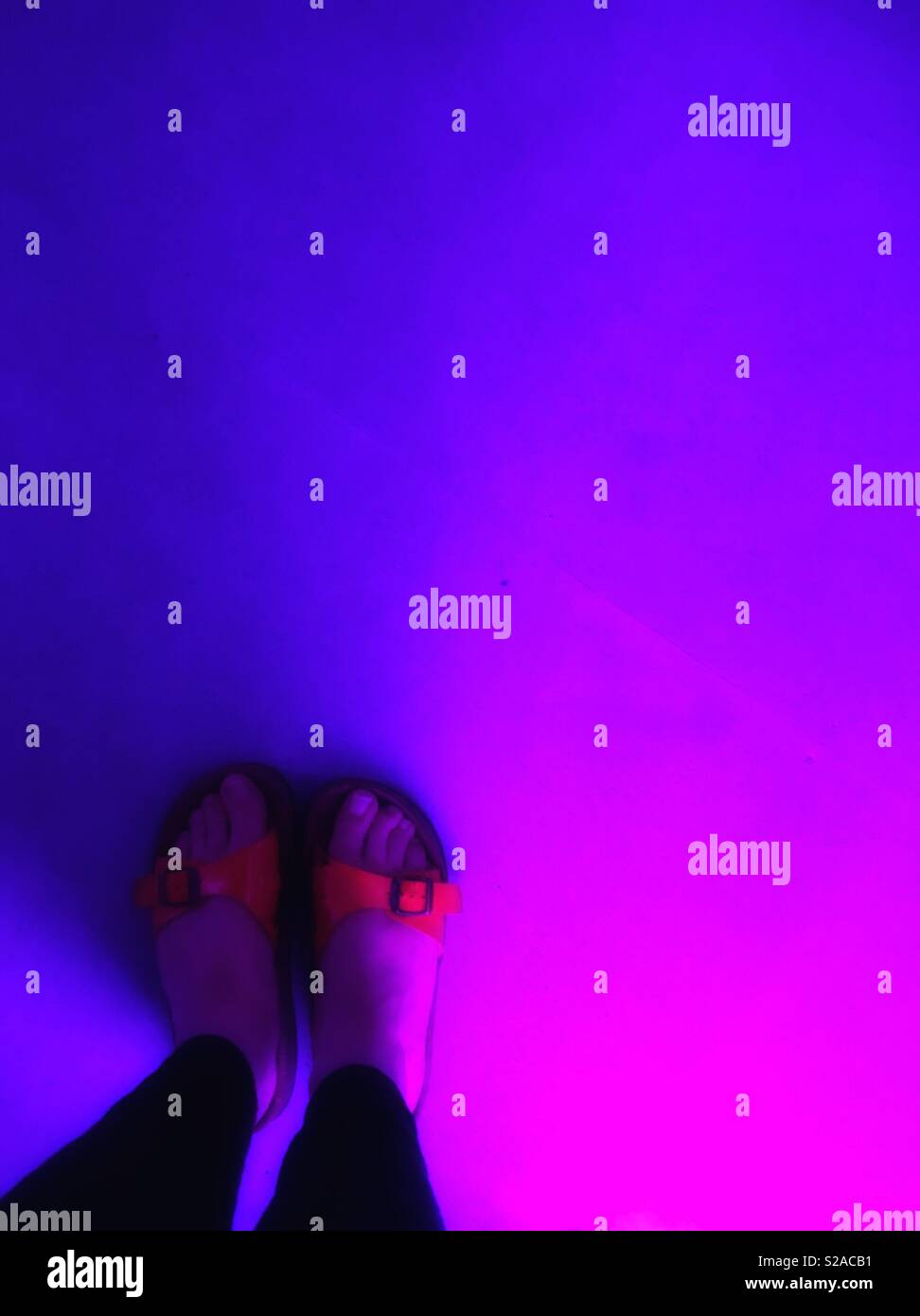 Il punto di vista della donna si affaccia su un paio di sandali arancioni con un'illuminazione rosa/viola sul pavimento Foto Stock