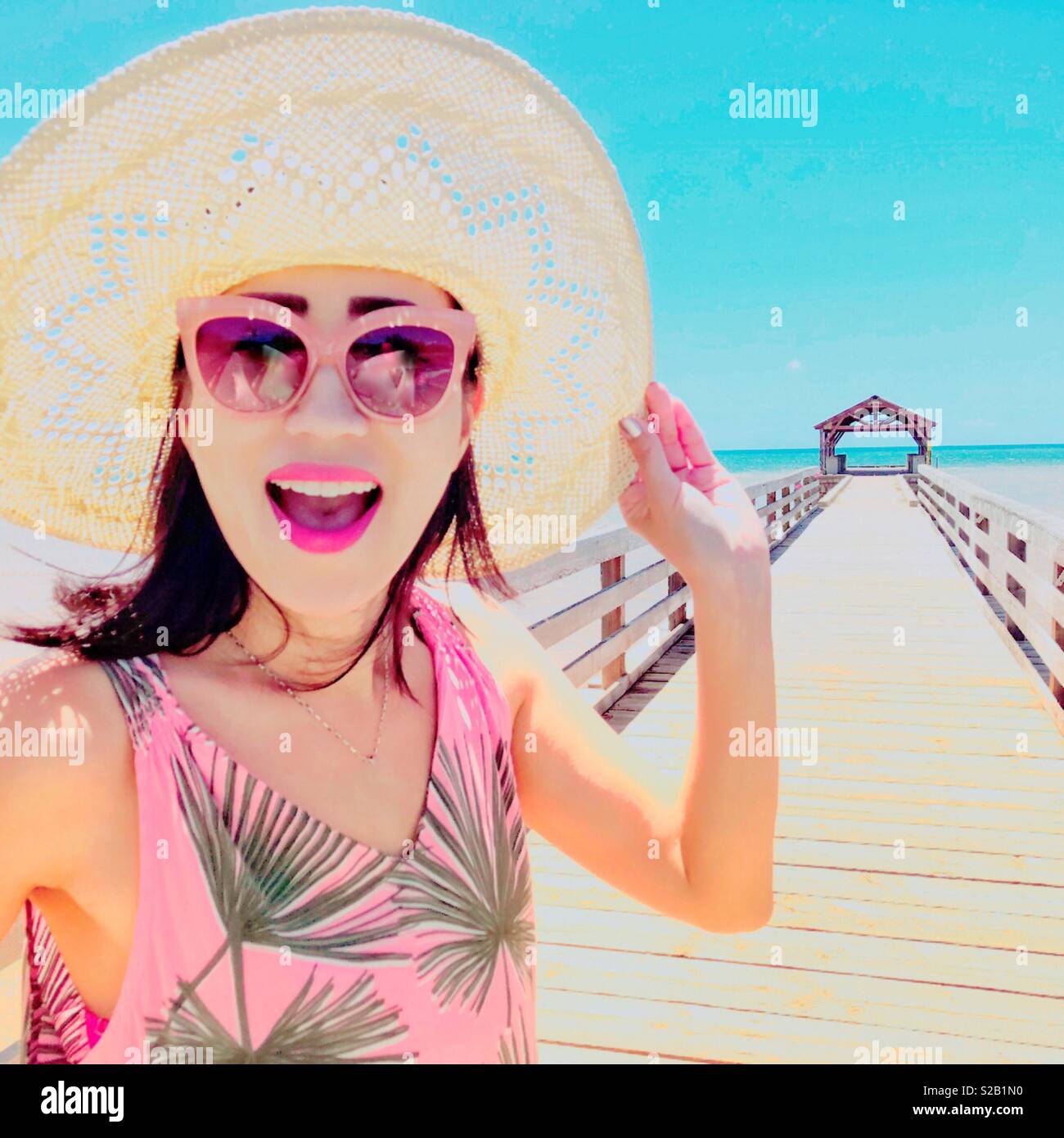 Vivace giovane donna in abito rosa, rosa occhiali da sole, rosa rossetto e cappello per il sole sorride per voi su un lungo molo che si affacciano sull'oceano tropicale, Waimea Hawaii Foto Stock