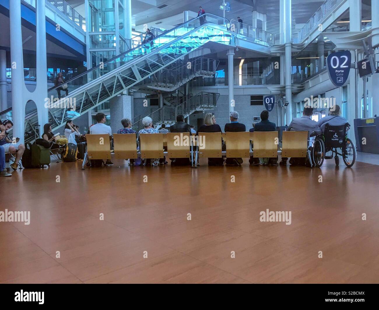 Passeggeri seduti al cancello dell'aeroporto di Porto, Portogallo. Interni moderni del terminal con ascensori in vetro, scale mobili e segnaletica per l'accesso con sedia a rotelle. - Immagine stock catturata con smartphone