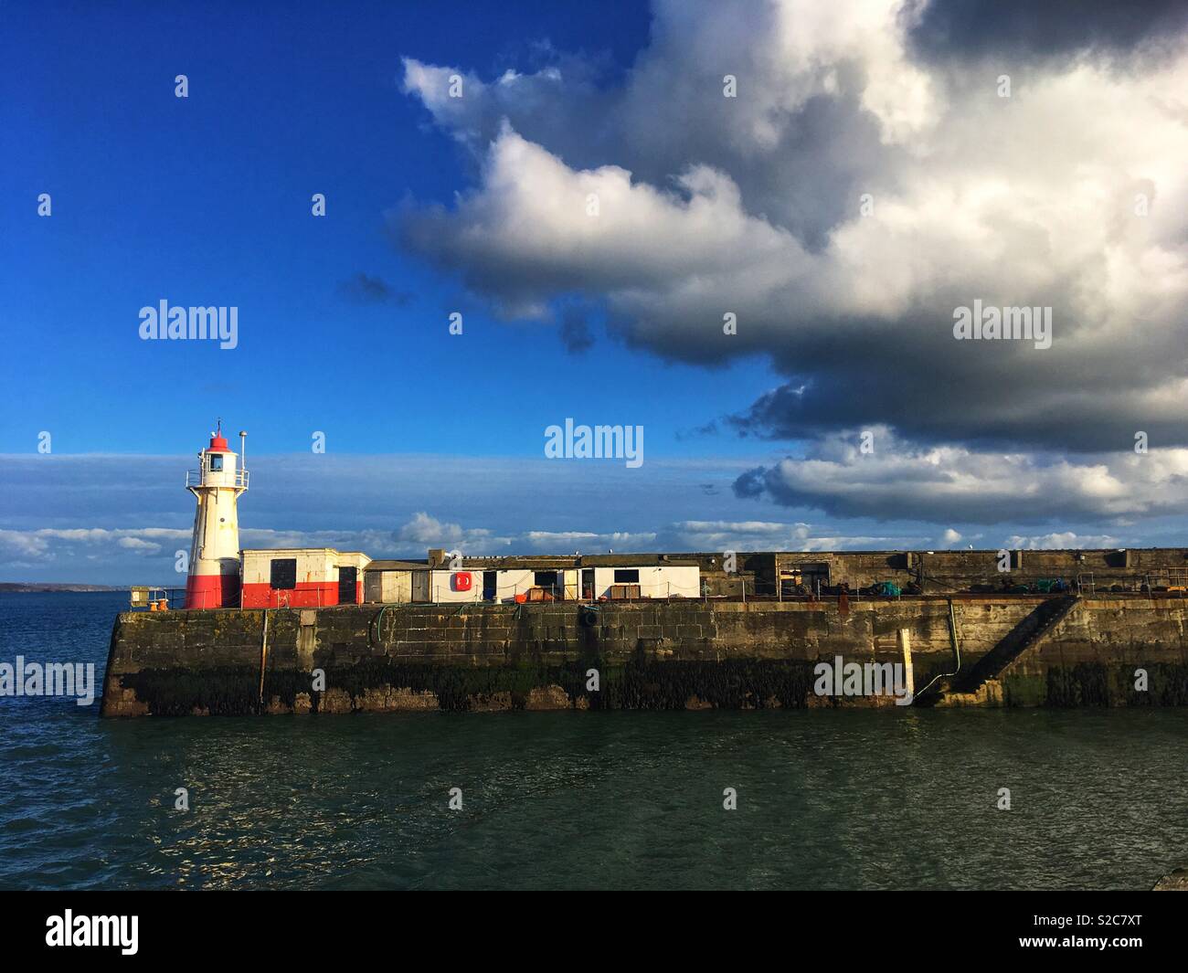 Faro ed edifici dipinti di rosso e bianco sul bordo del Porto di Newlyn Cornwall, cielo blu con un grande bianco e grigio cloud Foto Stock