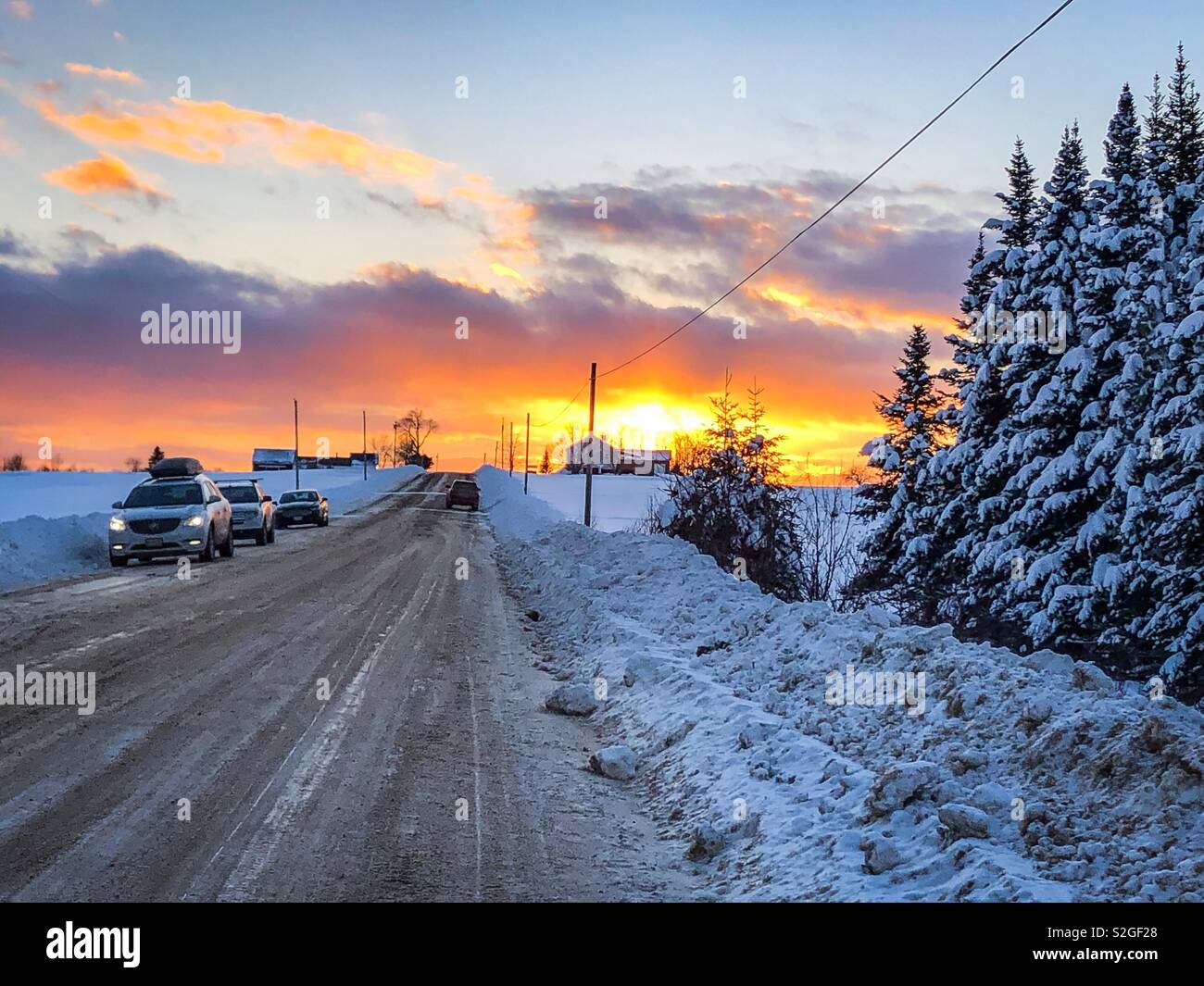 Una coperta di neve strada rurale al tramonto. Le automobili sono fermato sul lato della strada. Un agriturismo può essere visto in lontananza. Foto Stock