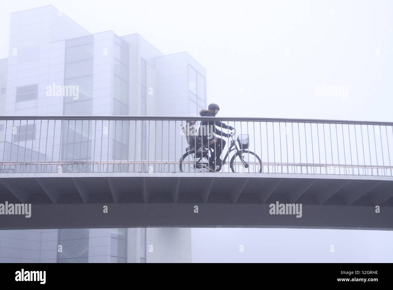 Una donna con un bambino sul sedile posteriore in bicicletta su un ponte in bicicletta nella nebbia meteo Foto Stock