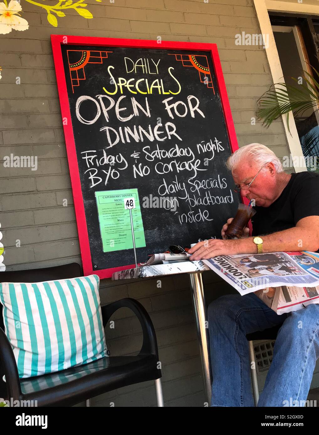 Un vecchio uomo seduto a un tavolo esterno di un coffee shop in Yamba, NSW, Australia. Egli sta leggendo un giornale e bere una bevanda fredda con una cannuccia, in quanto si tratta di una calda giornata estiva in Australia. Foto Stock