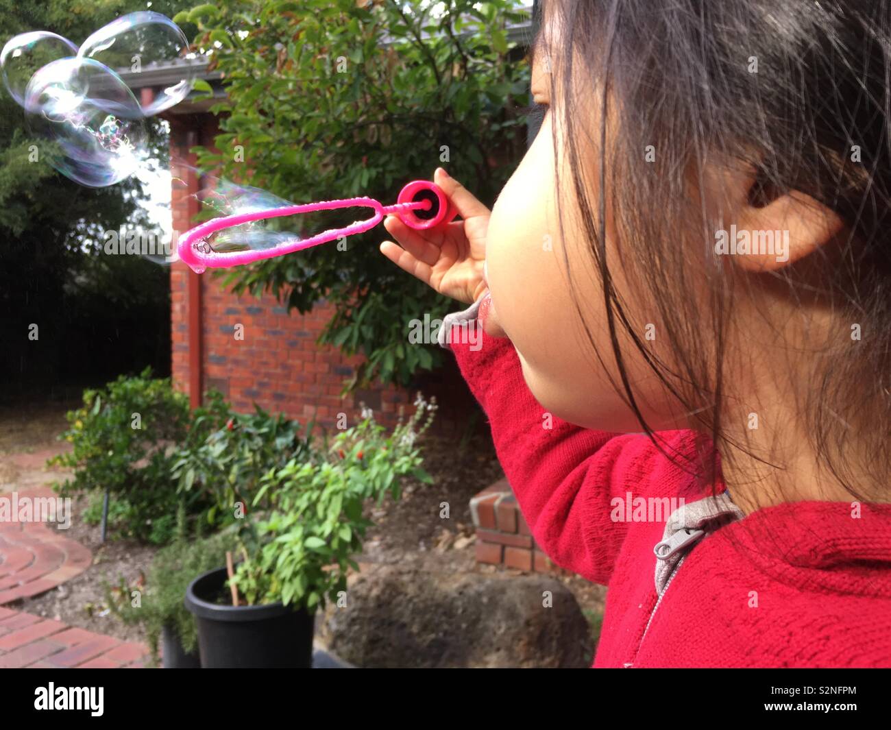 Un giovane multi etnico bambina indossa un cappuccio rosso il ponticello è soffiare bolle in un cortile. La stagione autunnale. Foto Stock