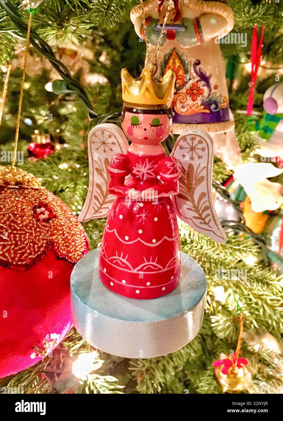 Angelo in legno ornamento in abito rosso che indossa corona d'oro pregando su albero di Natale è circondato da altri ornamenti e aghi di pino - Immagine stock catturata con smartphone