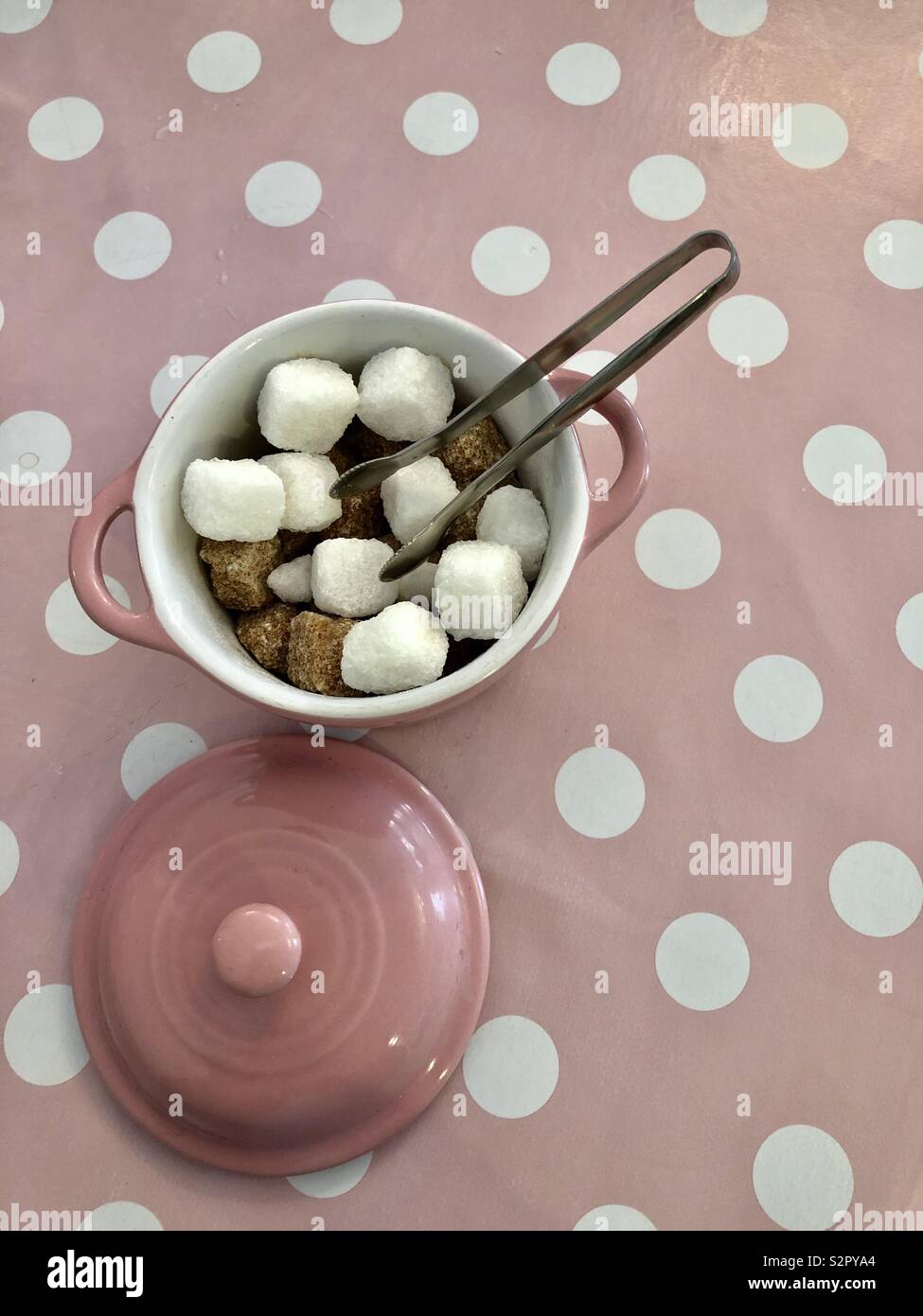 Marrone e bianco cubetti di zucchero e pinze da zucchero in una ciotola rosa su una rosa e bianco sfondo maculato Foto Stock