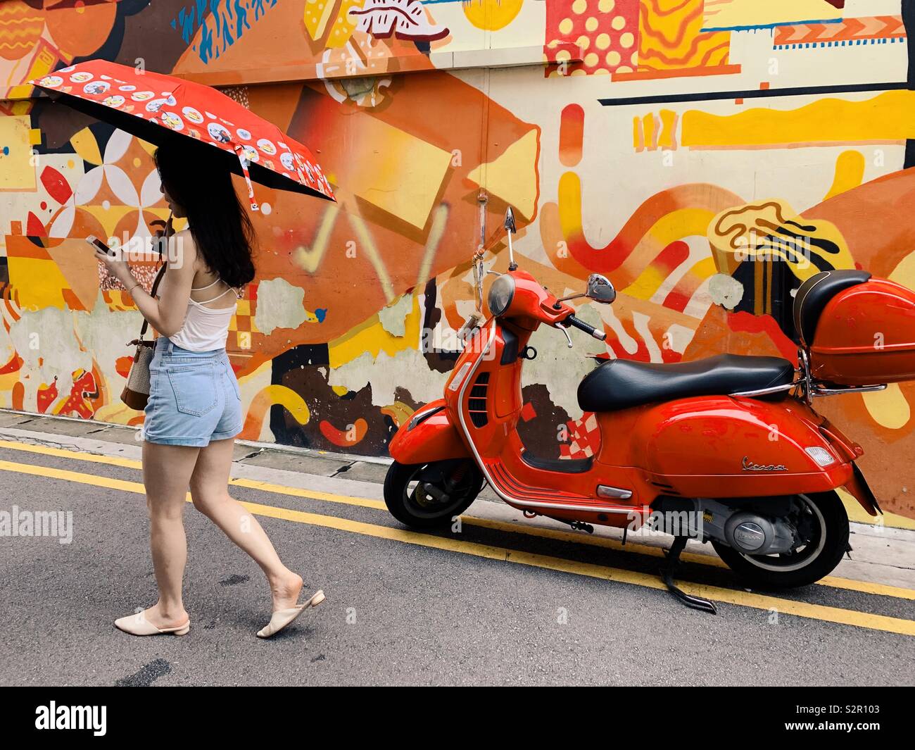Donna con ombrellone guardando telefono passato a piedi scooter e arte di strada sul muro a Haji Lane in Singapore. Foto Stock