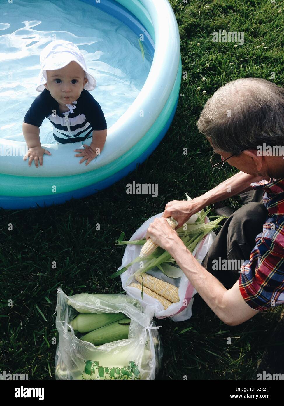 Bambino in un striped costume e cappello bianco si appoggia sul bordo di un  blu piscina gonfiabile a guardare il suo nonno vestito di reed plaid  manicotto corto shuck bucce di mais