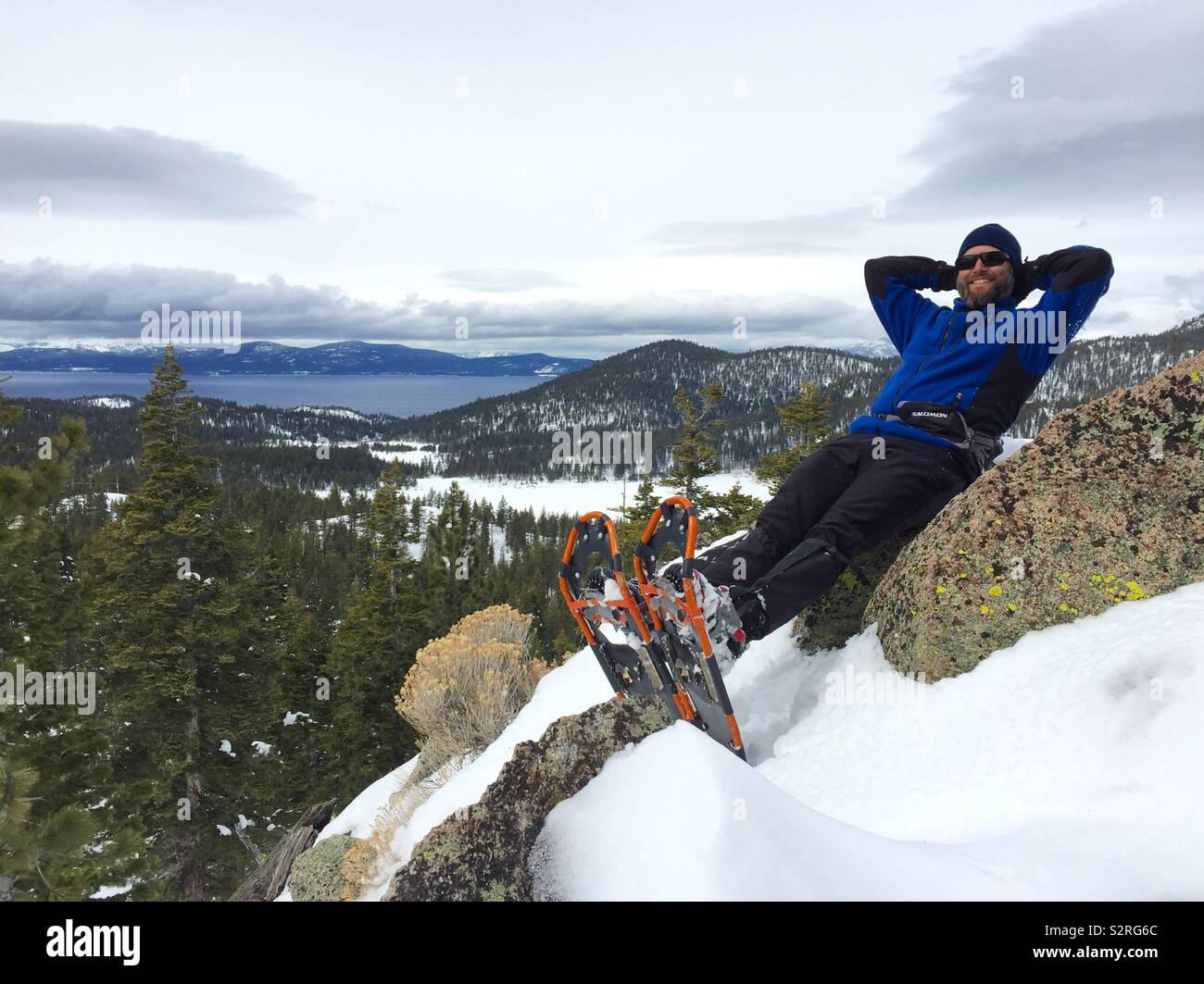 Un uomo con le racchette da neve appoggia la schiena su una roccia con il lago Tahoe in background. Foto Stock