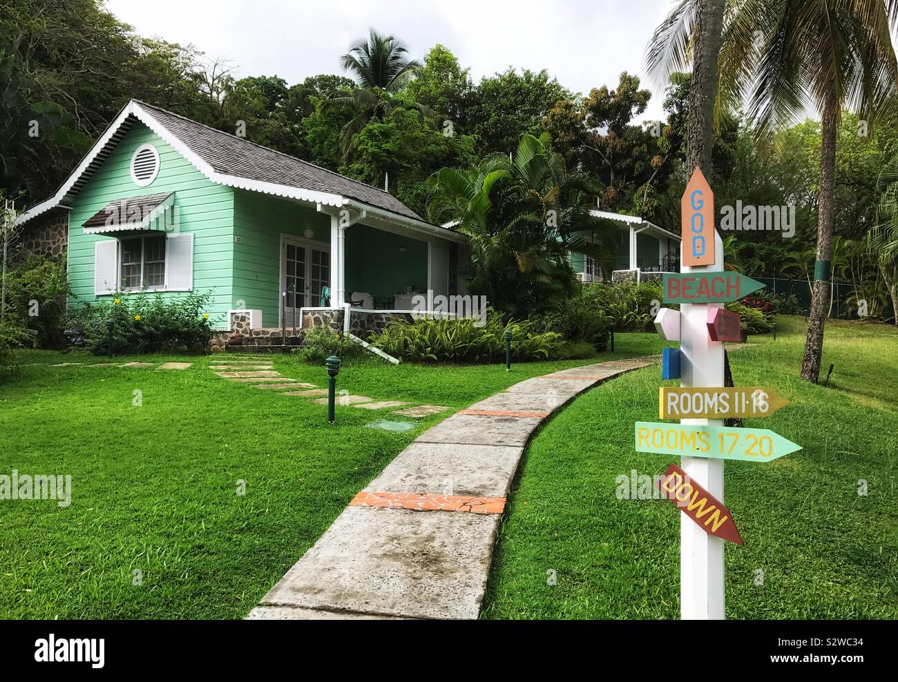 Giardino tropicale con un percorso e un cartello direzionale che conduce ad un verde Gingerbread Cottage con un portico anteriore. East winds Inn St.Lucia, West Indies Foto Stock