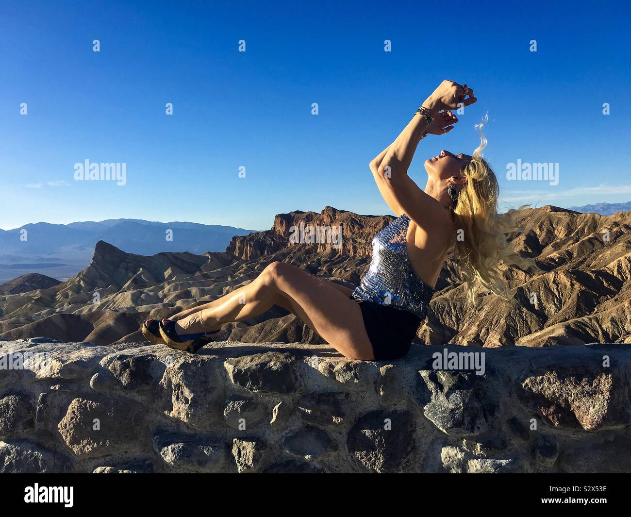 Modello bionda in argento tank top e pantaloncini nero seduto su una mensola che si affaccia Zabriskie Point nella Valle della Morte, una ciocca di capelli verso l'alto flottante per il cielo. Foto Stock