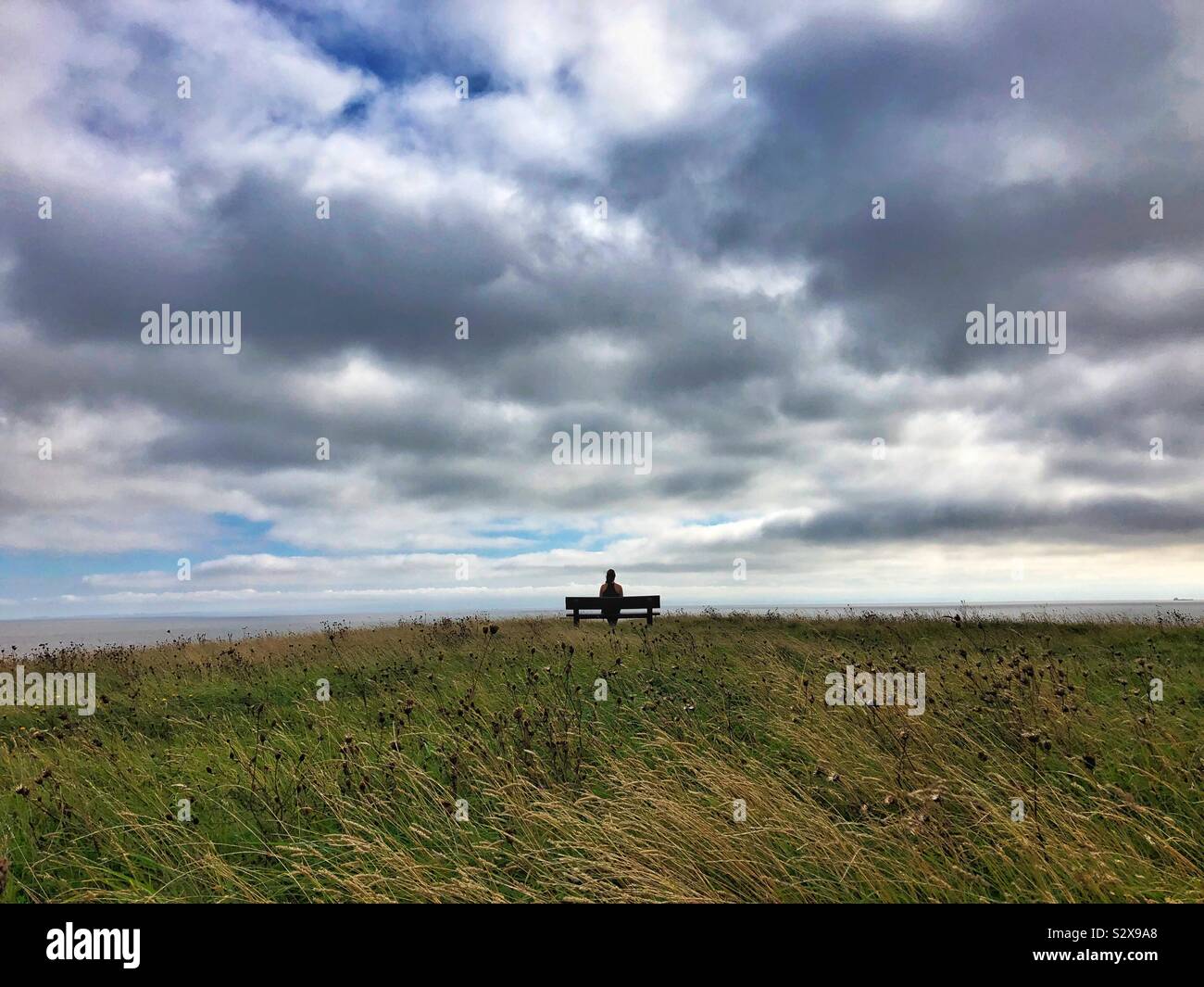 Giovane donna seduta su una panchina guardando il mare e il cielo. Foto Stock