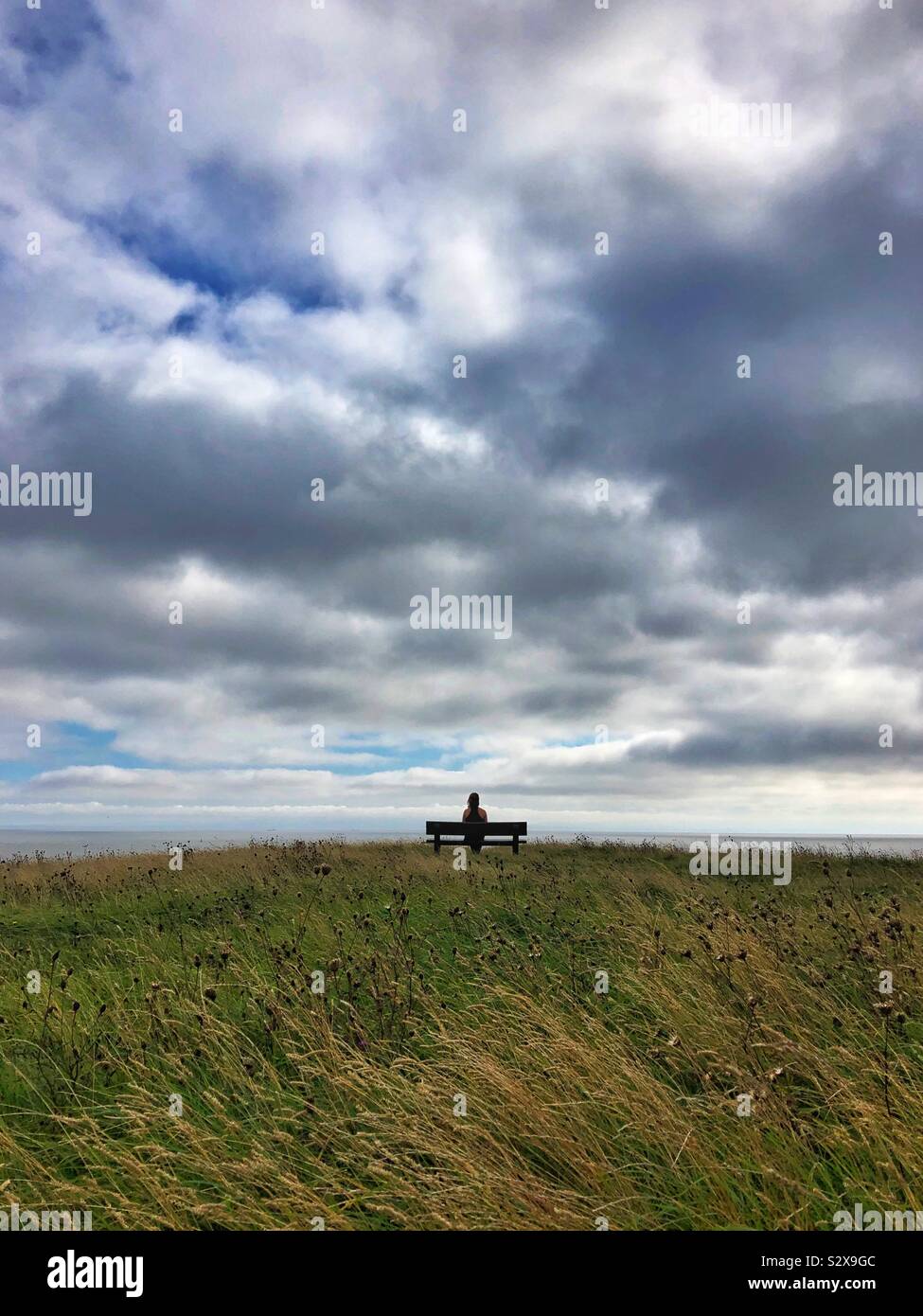 Giovane donna seduta su una panchina guardando il mare e il cielo. Foto Stock