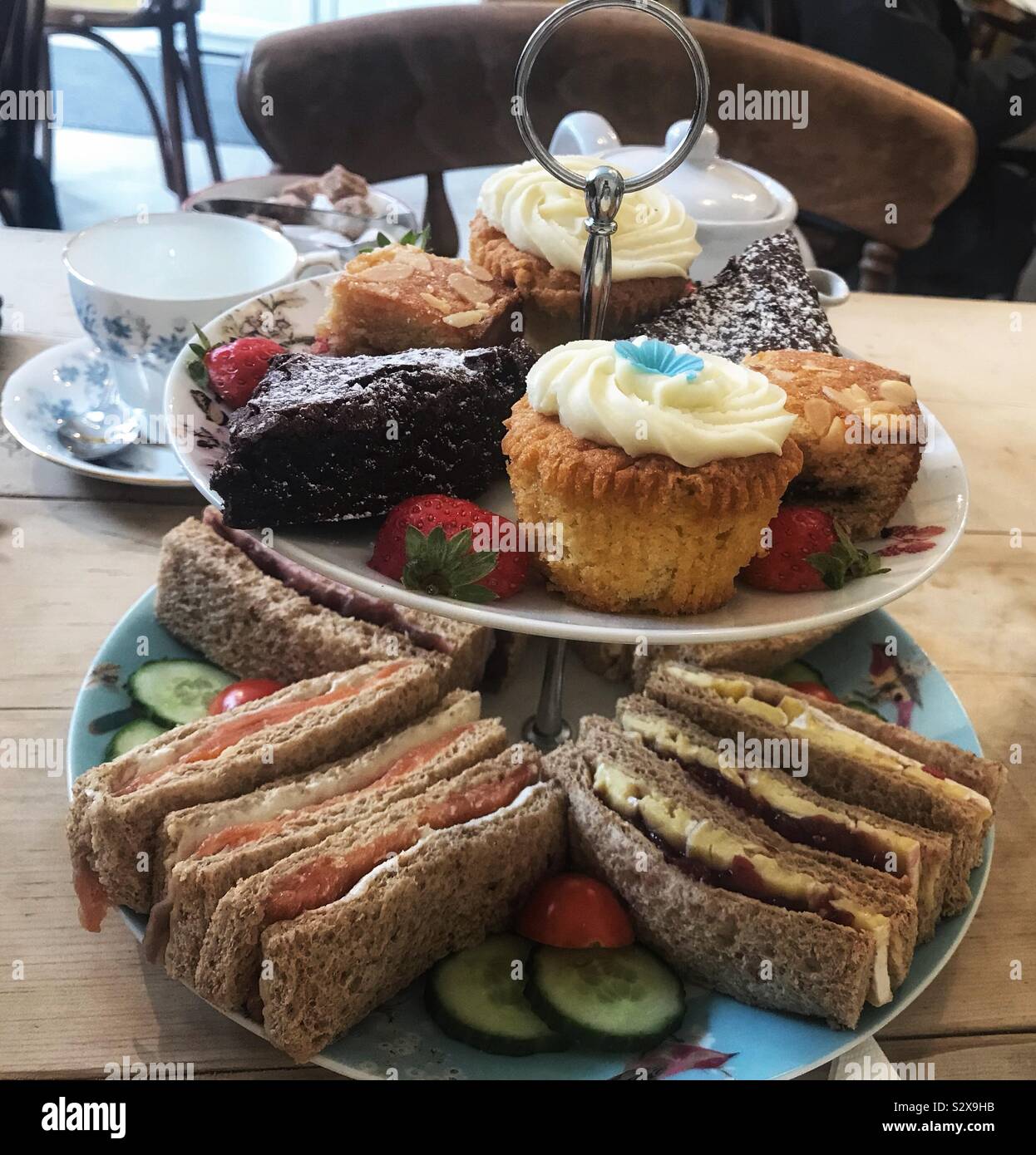 Il tè del pomeriggio su due Tier stand con una selezione di panini e torte su un tavolo di legno con una tazza e piattino Foto Stock