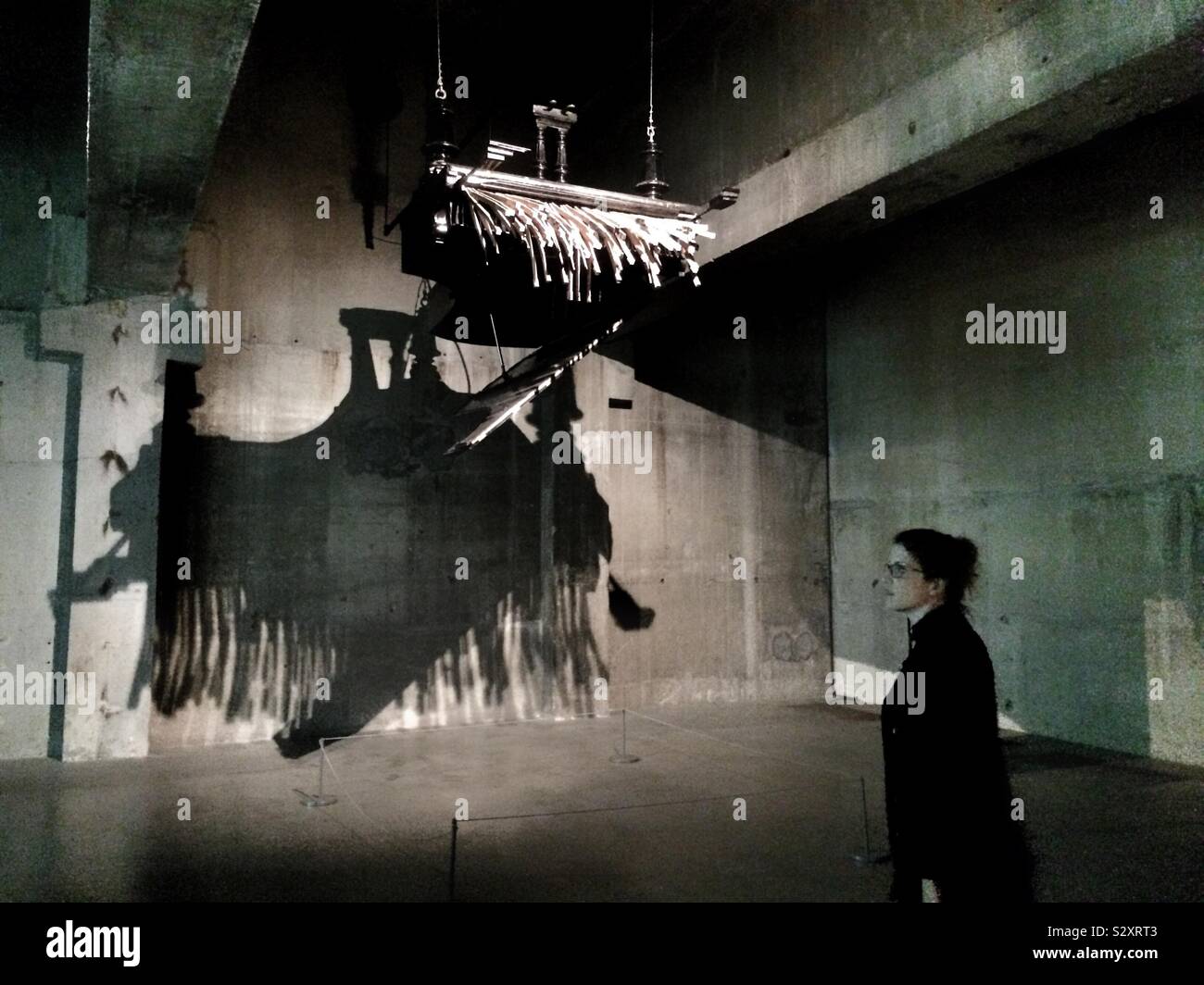 Una donna che guarda la scultura nella Tate Modern building - un pianoforte sospesa a testa in giù dal soffitto getta un ombra sul muro dietro - Immagine stock catturata con smartphone