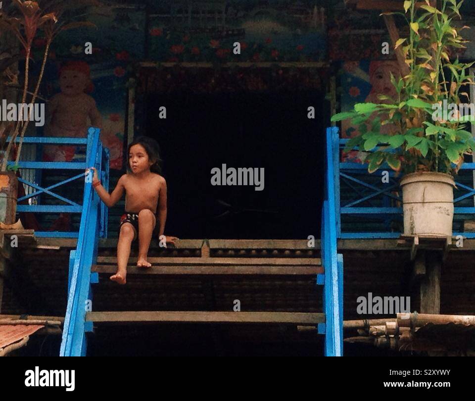 Un ragazzo cambogiano aspettando pazientemente sui gradini della sua casa su palafitte. Foto Stock