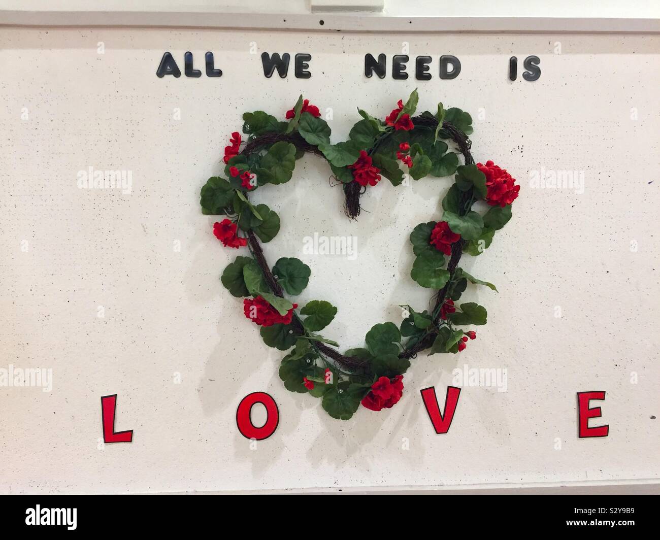 Tutto ciò di cui abbiamo bisogno è amore. Una corona floreale a forma stilizzata di cuore su un muro esprime il desiderio collettivo e universale di amore. Concetti: Noi, il nostro bisogno, l'amore è tutto ciò di cui avete bisogno. Nei nostri cuori. - Immagine stock catturata con smartphone
