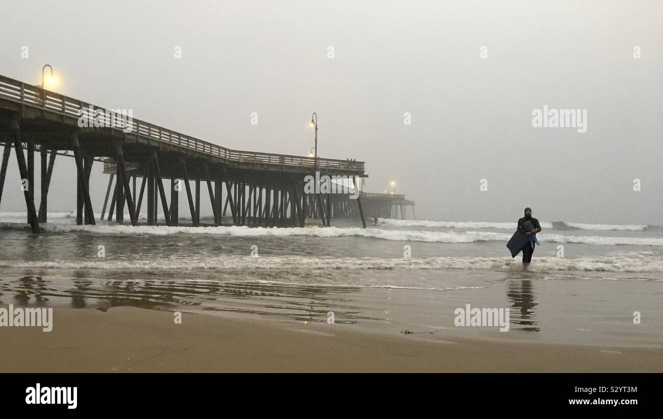 PISMO Beach, CA, OTT 2019: surfer in nero muta passeggiate fuori dell Oceano Pacifico accanto al molo, su un nebbioso pomeriggio tardi Foto Stock