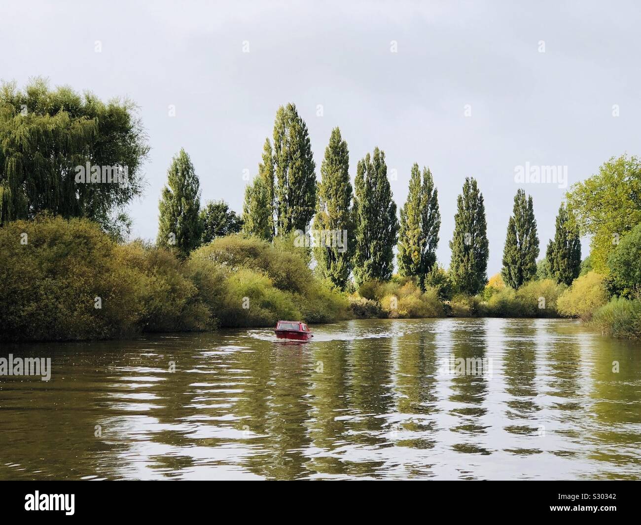 Rosso barca voce verso un fiume con una linea di alberi di pioppo riflessa Foto Stock