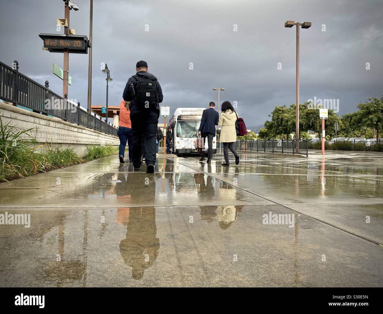 GLENDALE, CA, NOV 2019: i viaggiatori a piedi verso gli autobus con riflessi nelle pozze su un coperto, giornata piovosa a Glendale Metrolink Station - Immagine stock catturata con smartphone