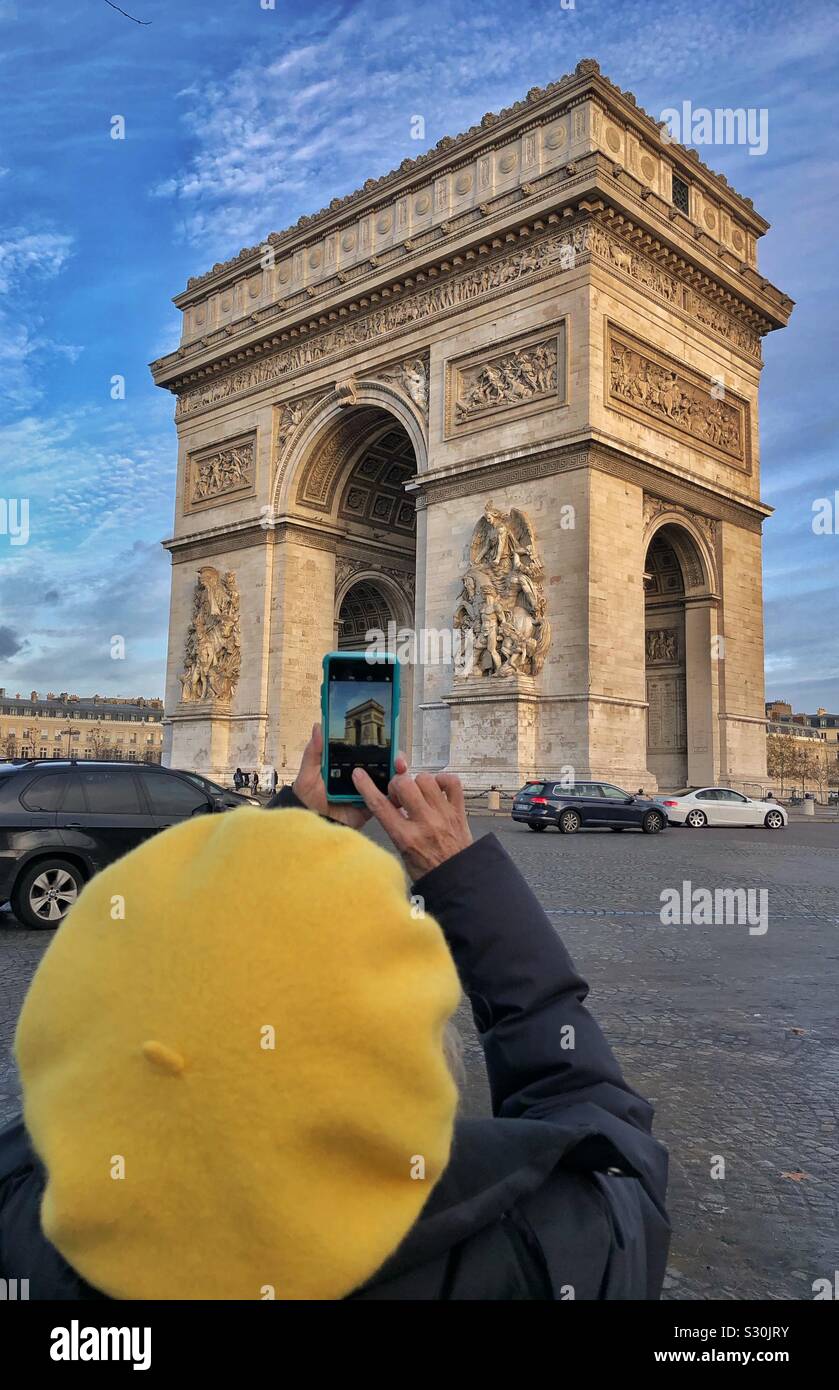 Un turista in una berta gialla scatta una foto dell'Arco di Trionfo a Parigi, Francia. L'immagine di Arch è visibile sullo schermo dell'iPhone. Foto Stock