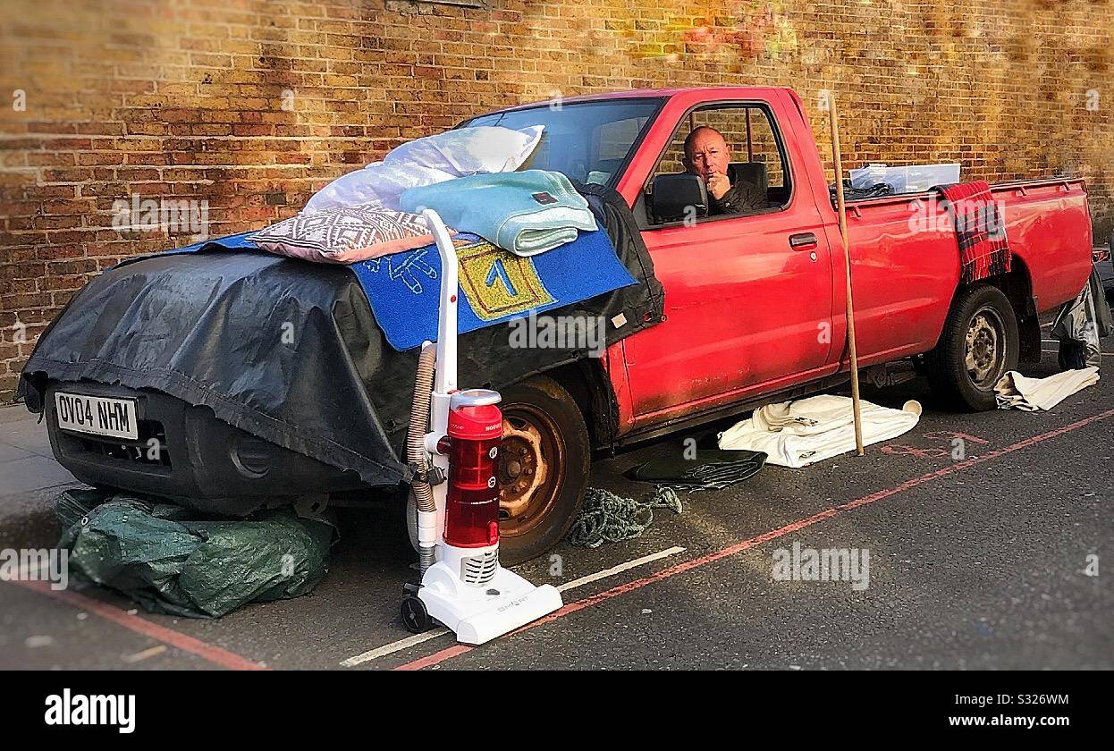 Un commerciante di mercato di strada seduto nel suo furgone rosso del pickup coperto e circondato da vari articoli per la vendita a Portobello Road, Londra Foto Stock