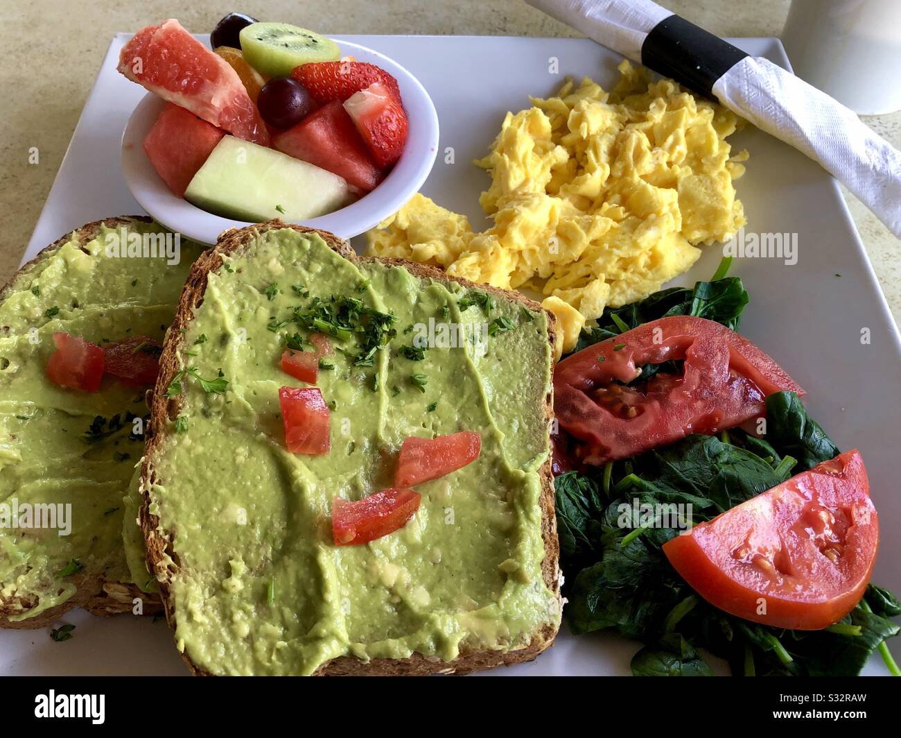 Piatto di cibi per la colazione: Toast all'avocado, uova strapazzate, frutta, spinaci e pomodoro. - Immagine stock catturata con smartphone