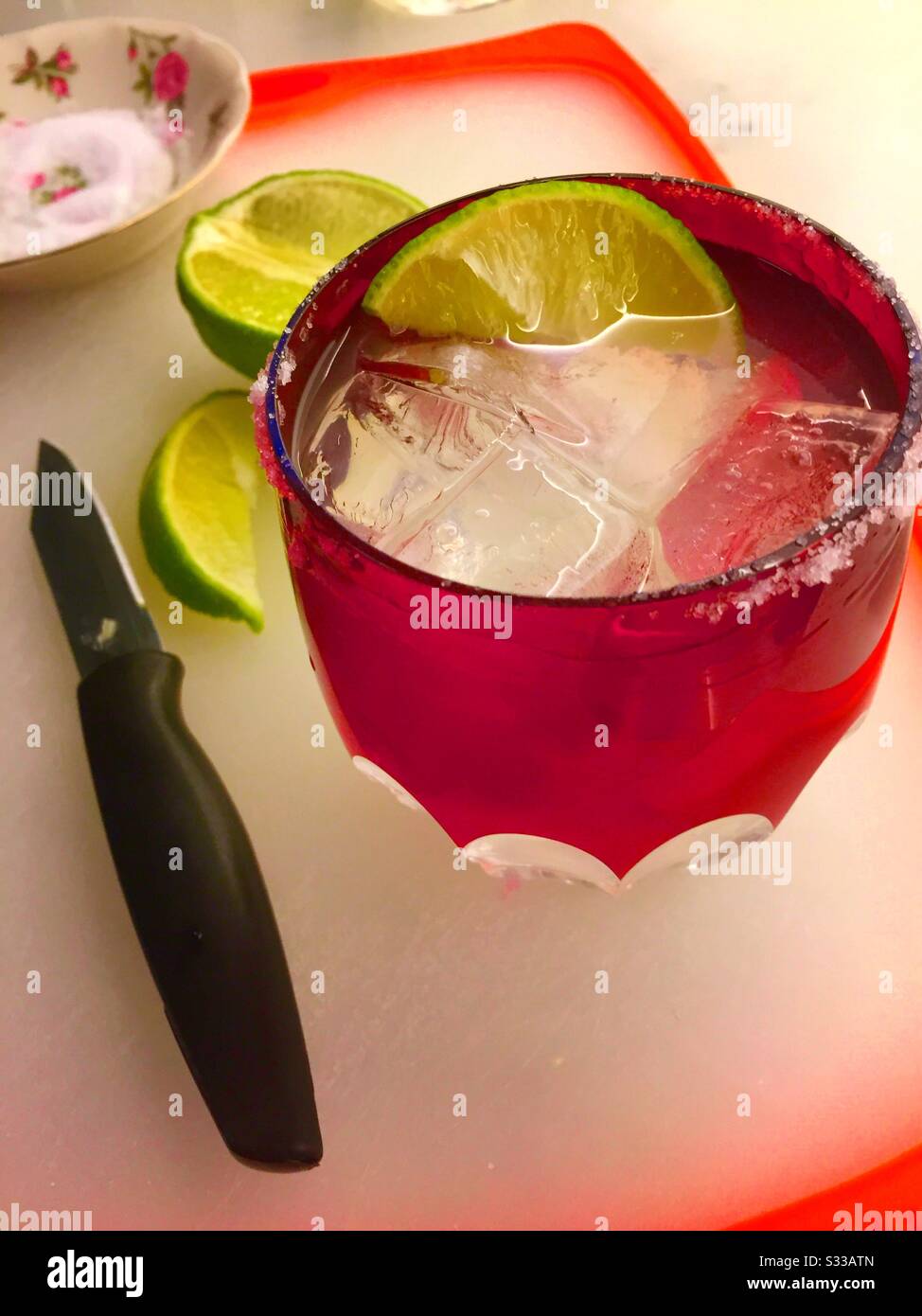 Margarita sulle rocce in un vetro di cristallo rosso con bordo salato con coltello sale e lime su un tagliere - Immagine stock catturata con smartphone Margarita sulle rocce in un vetro di cristallo rosso con bordo salato con coltello sale e lime su un tagliere - Immagine stock catturata con smartphone