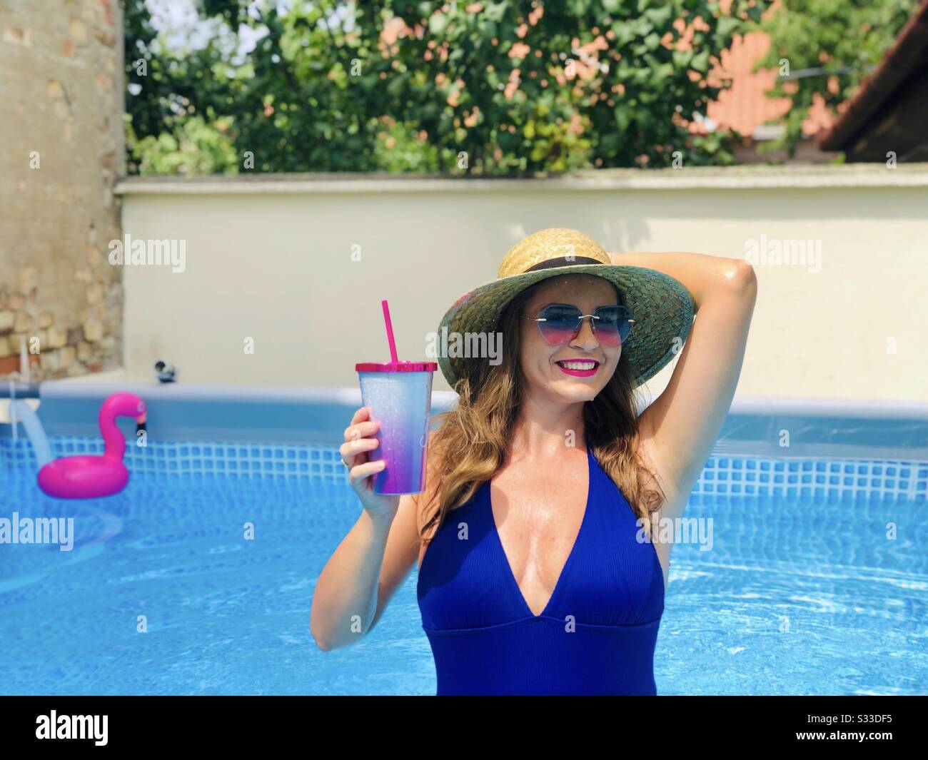 Donna che indossa bikini blu, occhiali da sole e cappello di paglia, tenendo un drink in piscina Foto Stock