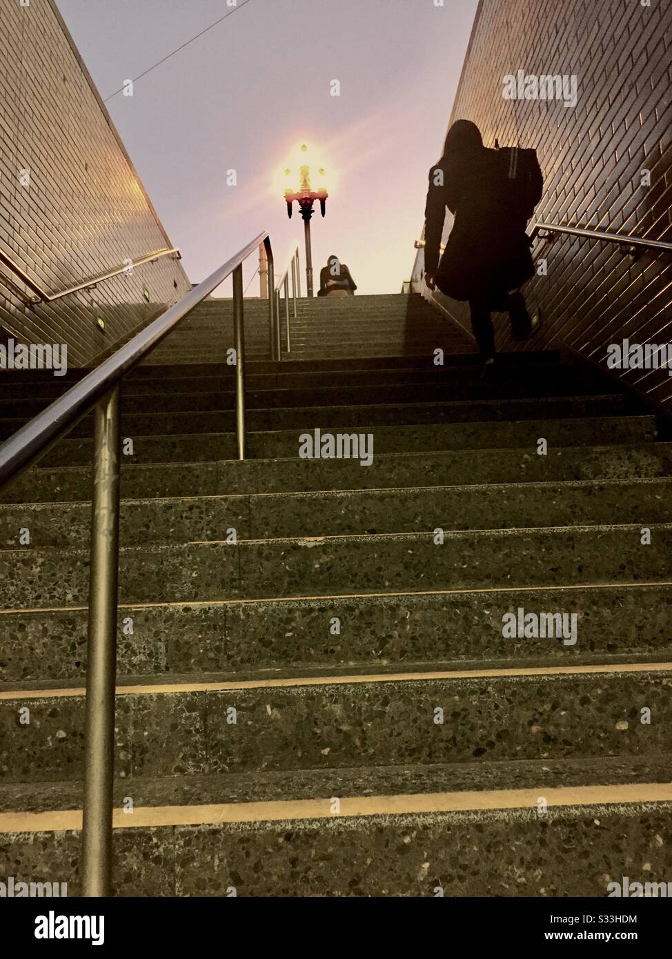 Due figure che salgono le scale dalla metropolitana sotterranea, metropolitana, o sistema di transito rapido al crepuscolo con ringhiera metallica e lampione. Reminiscenza delle scene dal joker. San Francisco, California. Foto Stock