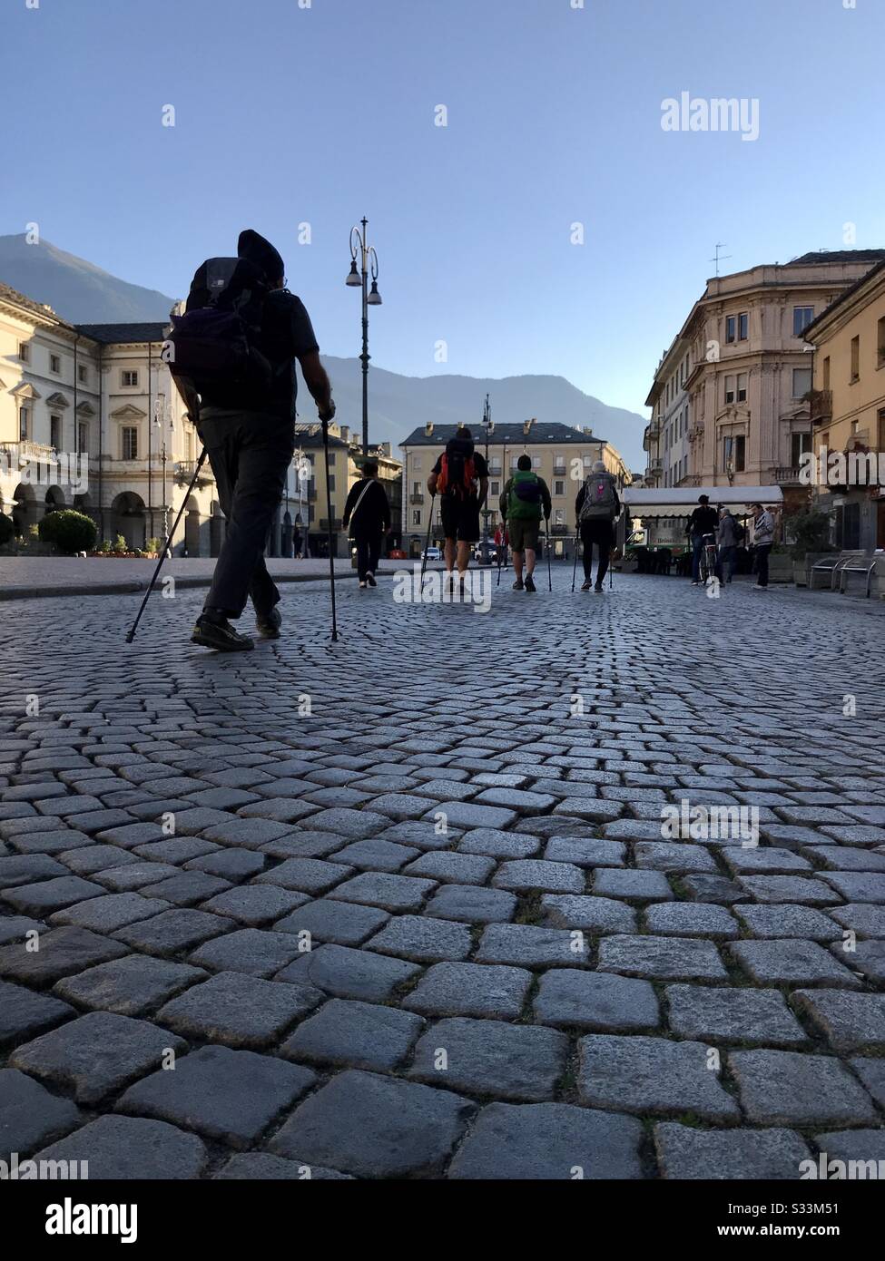 All'alba Via Francigena gli escursionisti Pellegrini camminano attraverso il villaggio di Aosta, Italia. Strade acciottolate, spalle di escursionisti e vecchi edifici in tonalità gialla e un cielo blu. Foto Stock
