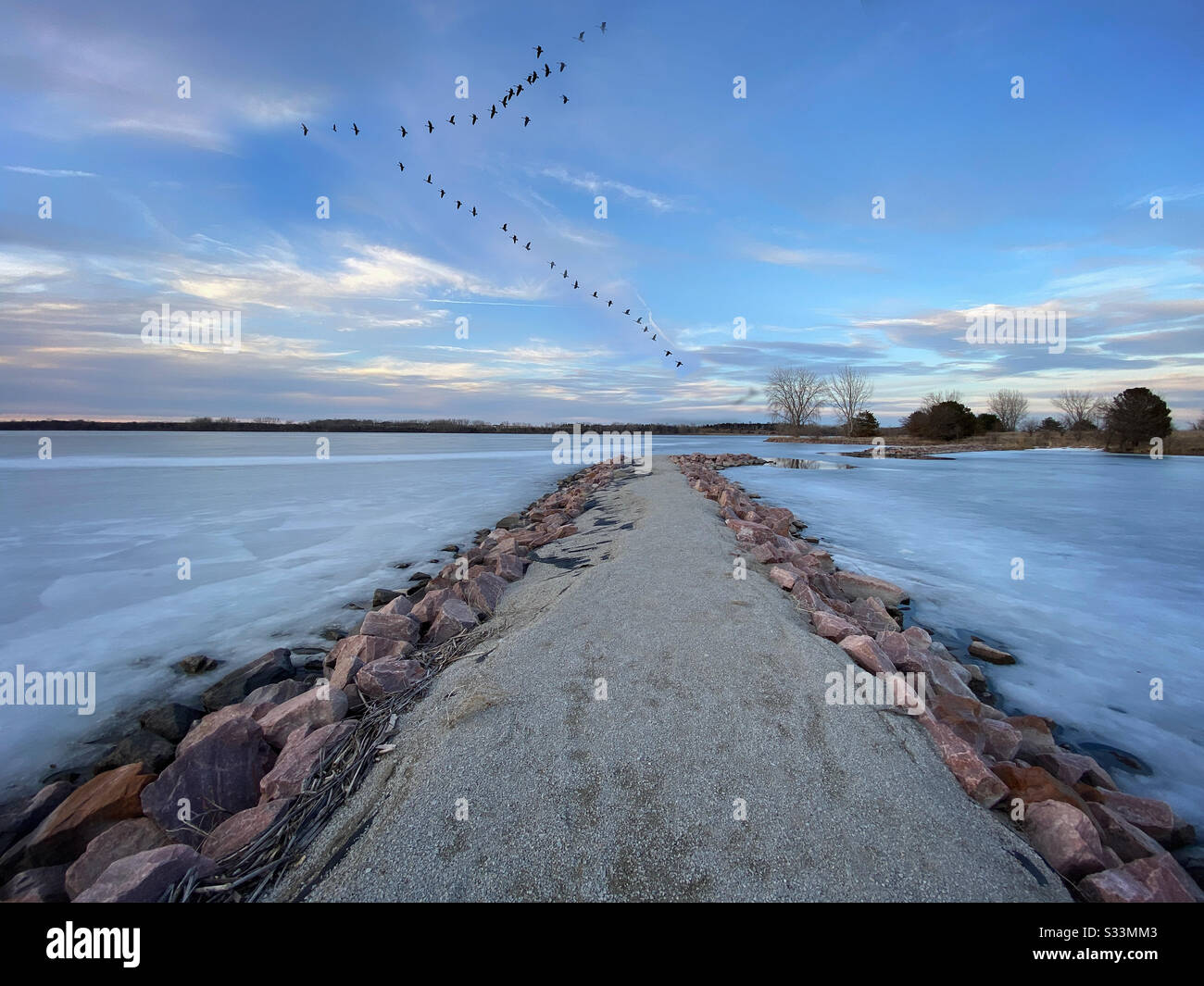 Un molo roccioso su un lago ghiacciato con oche volare in formazione a V sopra Foto Stock