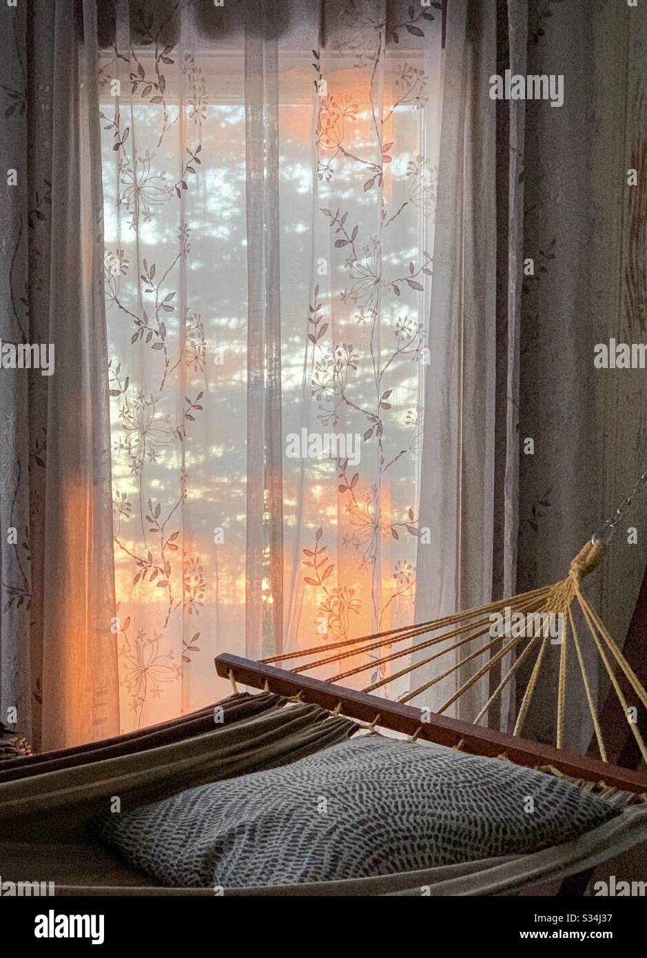 Un'amaca interna vi invita a riposare. La luce del sole del pomeriggio può essere vista attraverso le tende velate nella finestra. Foto Stock