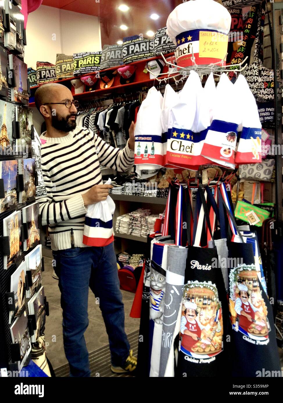 Uomo che fa shopping in un negozio di souvenir gastronomico guardando grembiuli a Saint-Louis, Parigi. - Immagine stock catturata con smartphone