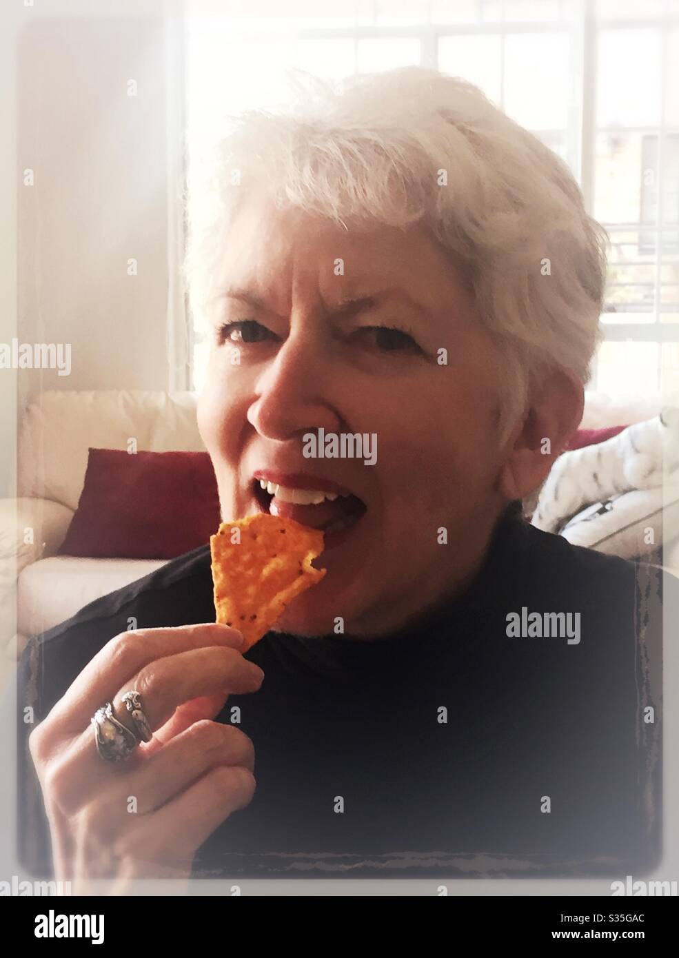 Donna anziana con capelli grigi che mangia un chip di Dorito per uno spuntino pomeridiano Foto Stock