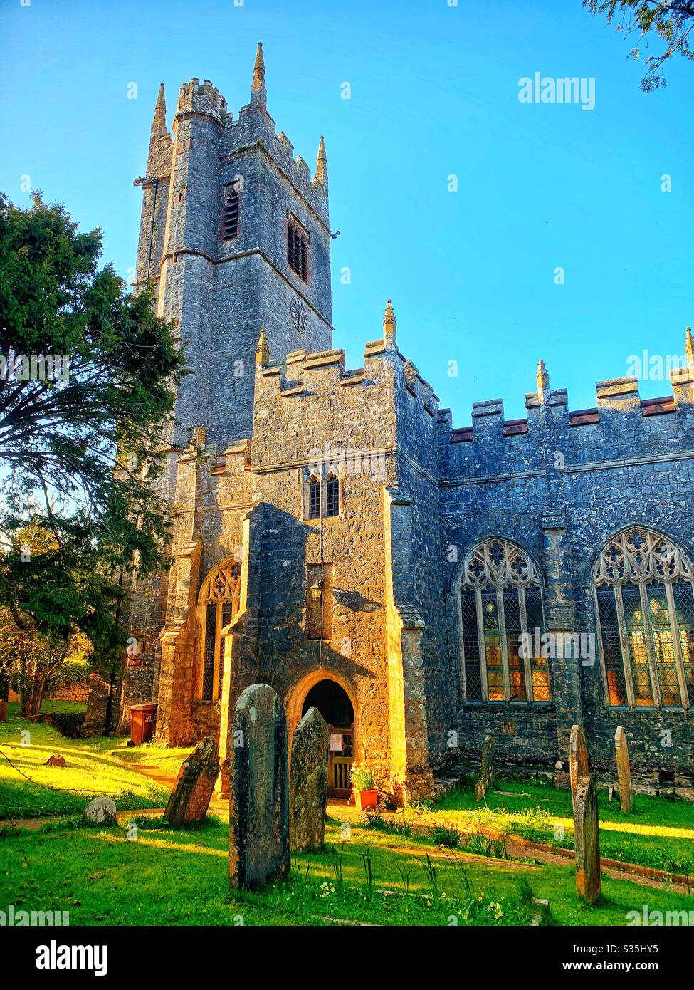 Una chiesa del villaggio Devon verso la fine della giornata con il sole dorato splendente sull'edificio e lapidi e alberi in primo piano. Foto Stock