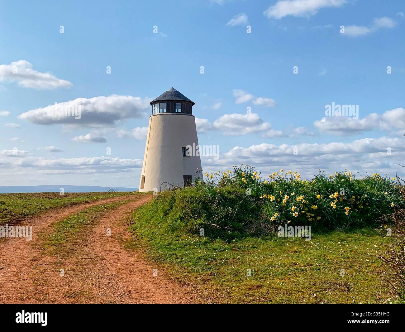 Vecchio edificio restaurato a vento, situato sulla cima di una verde collina con narcisi e un tortuoso sentiero fangoso per l'accesso. Cielo blu con nuvole. Foto Stock