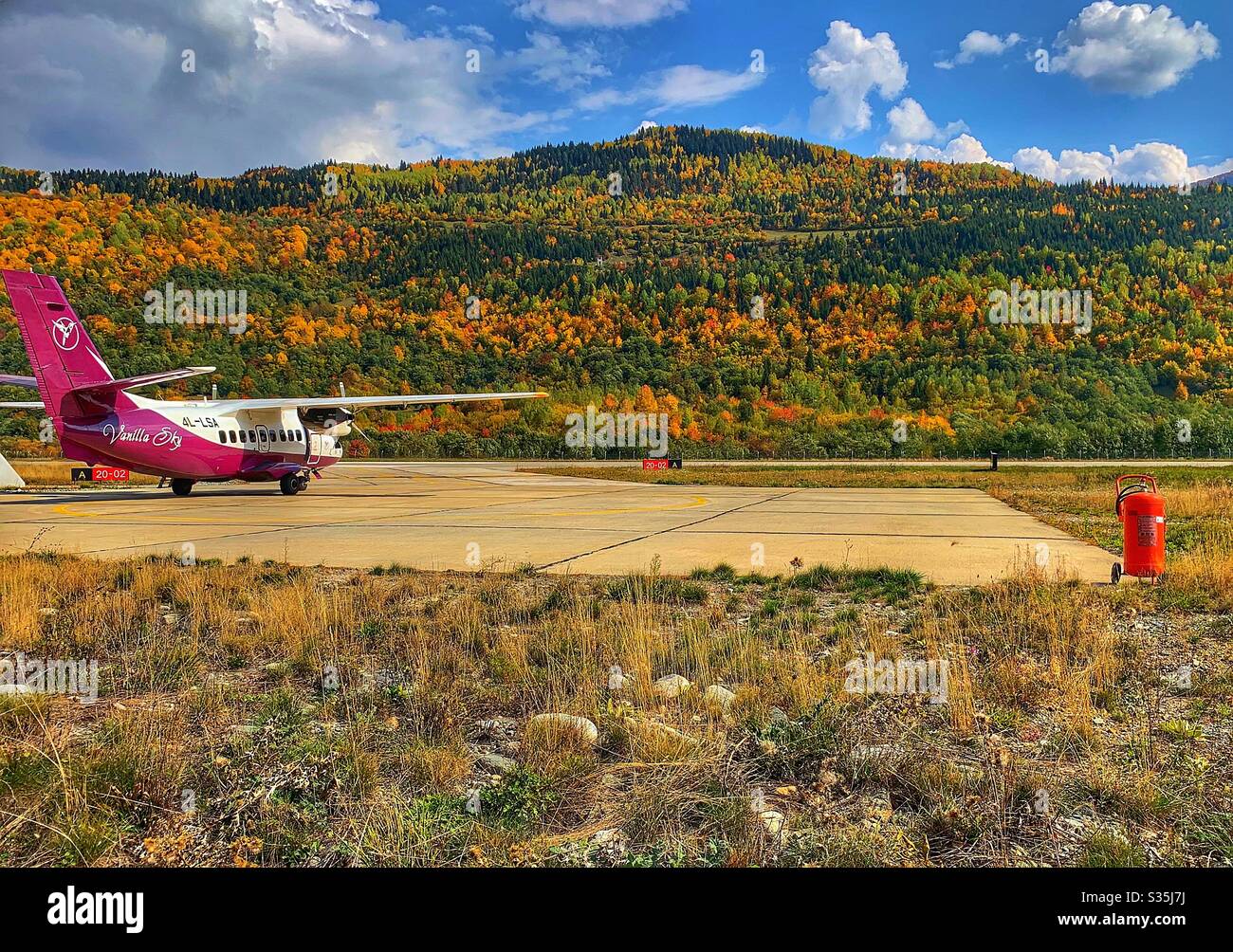 Un piccolo aeromobile gestito da Vanilla Sky è parcheggiato sul grembiule fuori dall'aeroporto di Mestia nelle montagne del Caucaso. Circondato da una foresta autunnale. Georgia. - Immagine stock catturata con smartphone