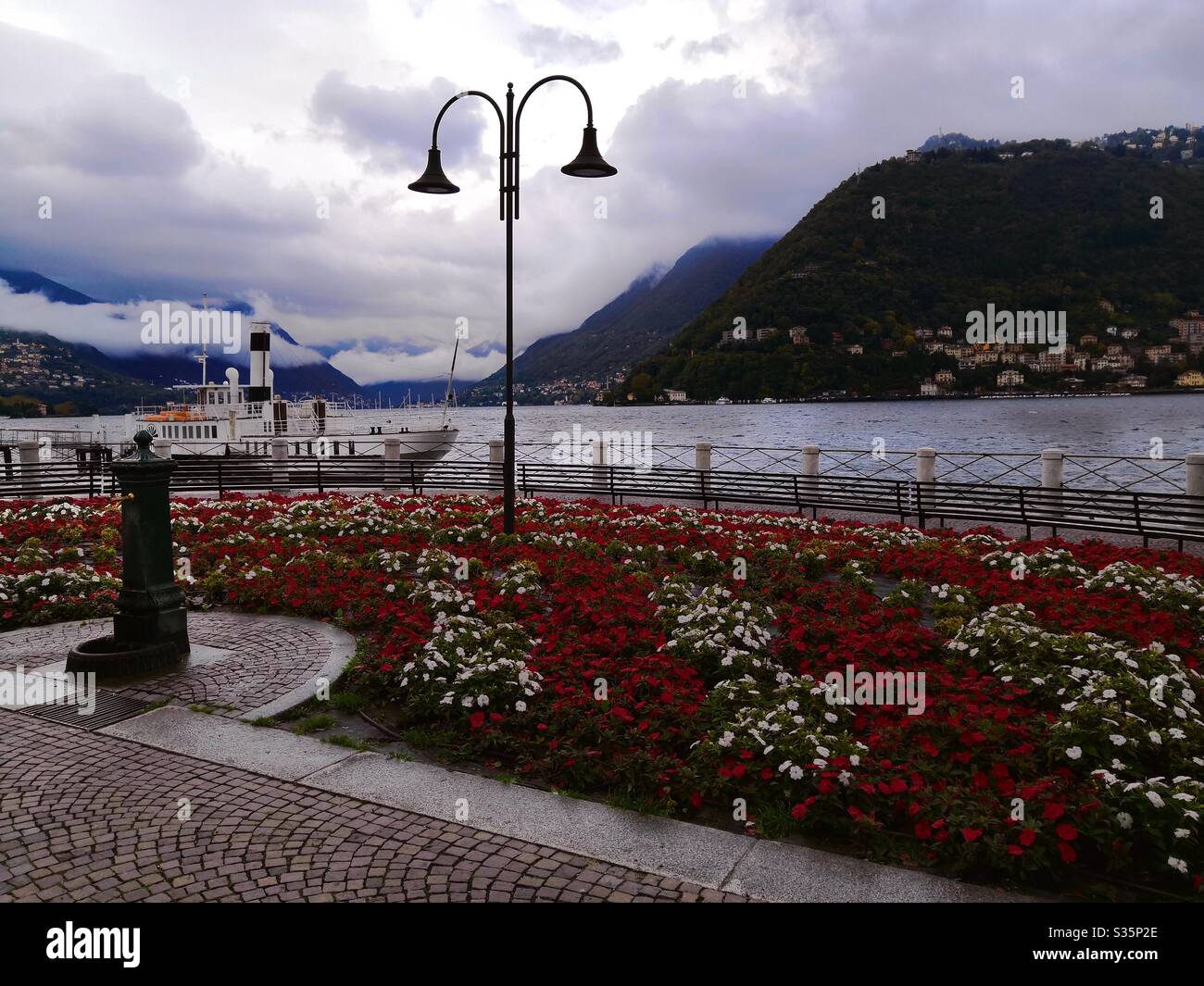 Piccolo giardino fiorito con fontana sul lato del Lago di Como in giornata nuvolosa Foto Stock