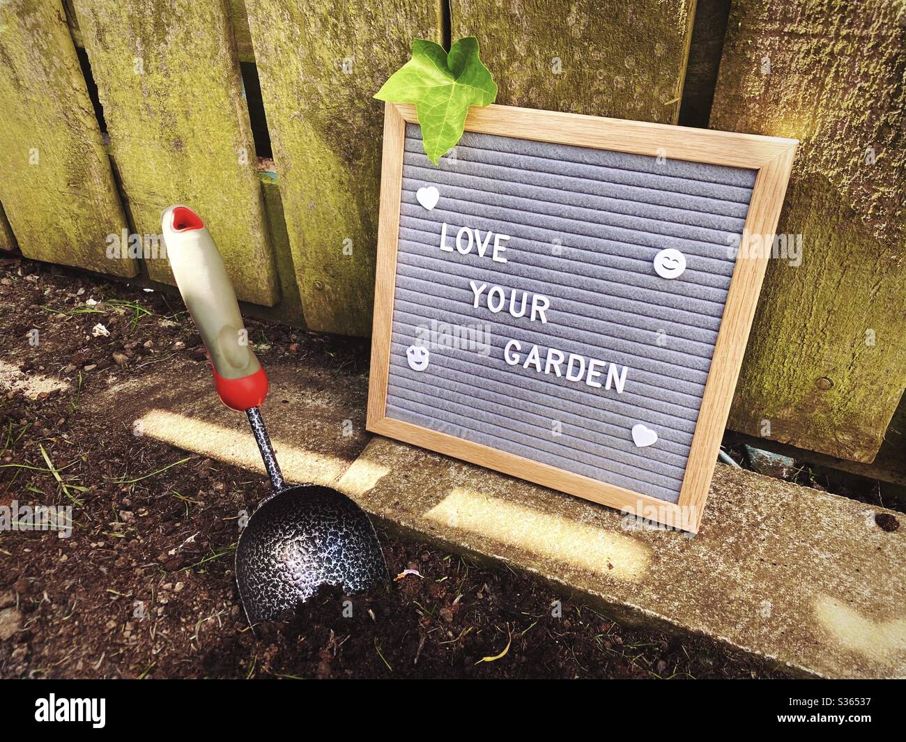 Ama il tuo giardino. Concetto di giardinaggio con bordo della lettera del feltro, attrezzi di giardinaggio, piccolo cazzone portatile. Lavorando ed erbando un bordo di suolo pronto per piante di primavera. Contro un recinto di legno intemperato. - Immagine stock catturata con smartphone