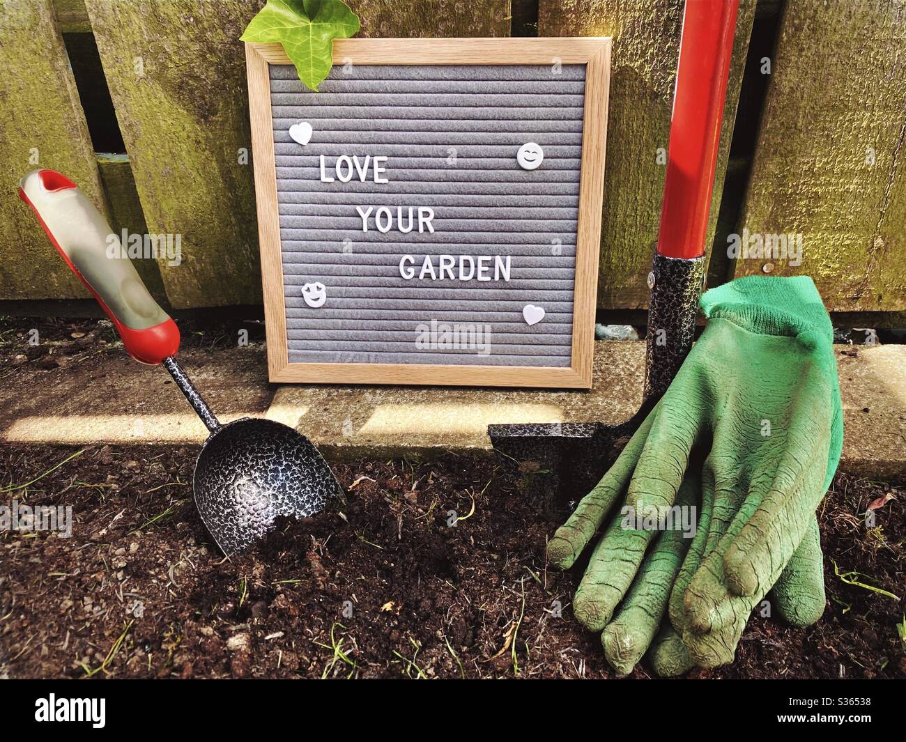 Ama il tuo giardino. Concetto di giardinaggio con bordo della lettera del feltro, attrezzi di giardinaggio, guanti sporchi. Lavorando ed erbando un bordo di suolo pronto per piante di primavera. Contro un recinto di legno intemperato. - Immagine stock catturata con smartphone