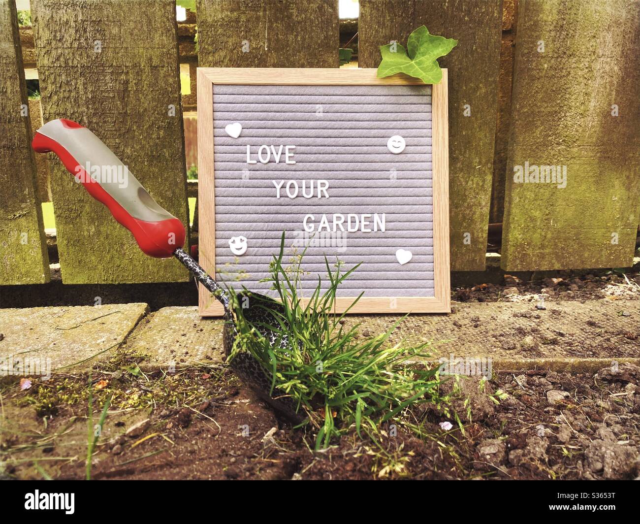 Ama il tuo giardino. Concetto di giardinaggio con bordo della lettera del feltro, attrezzi di giardinaggio, trowel della mano che scavano sulle erbacce. Lavorando ed erbando un bordo di suolo pronto per piante di primavera. Contro un recinto di legno intemperato. - Immagine stock catturata con smartphone