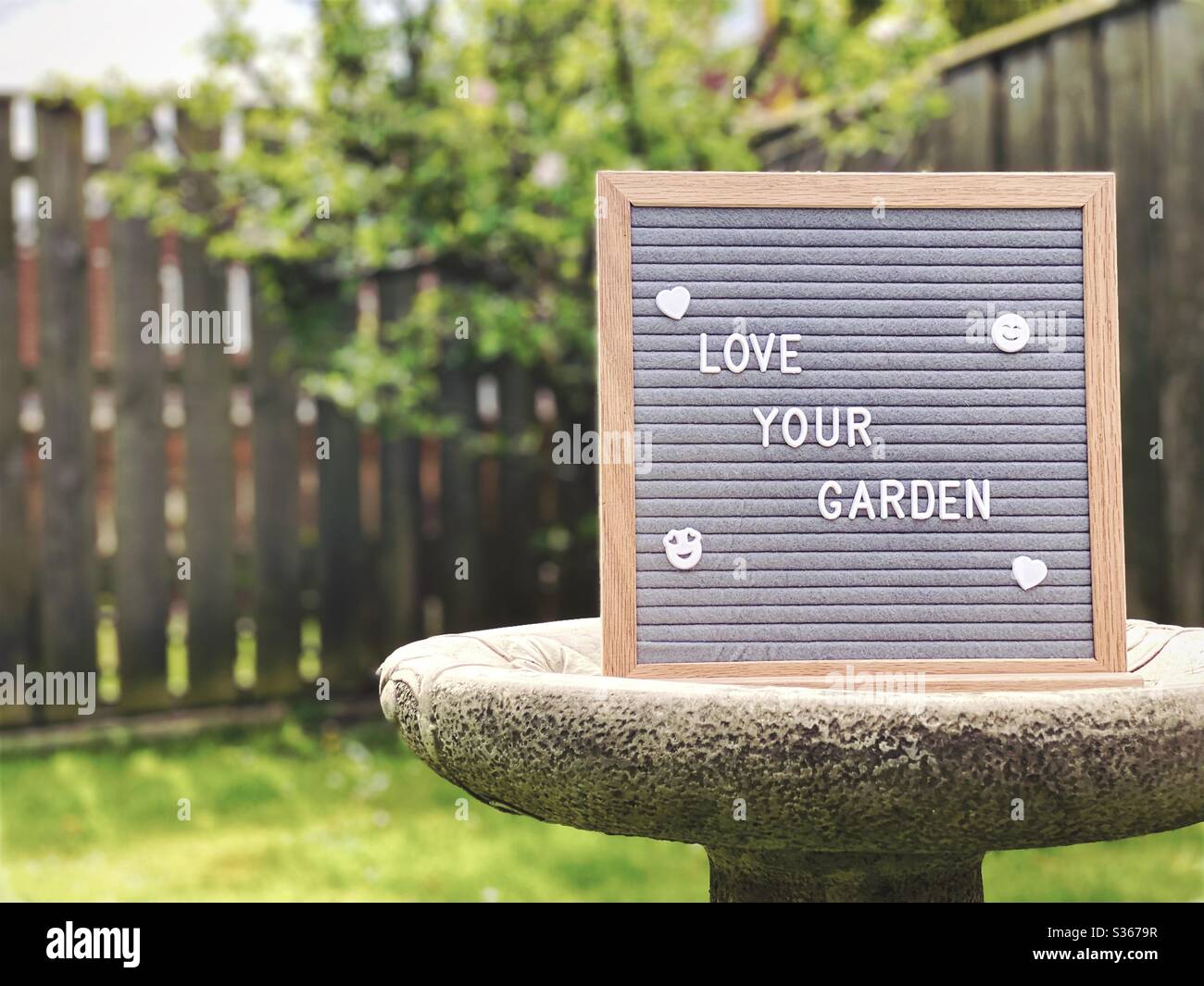 Amate il vostro giardino. Concetto di giardinaggio con bordo di messaggio feltro su un bagno di uccello nel mezzo di un giardino suburbano con l'albero di mela nana nello sfondo e spazio di copia. - Immagine stock catturata con smartphone