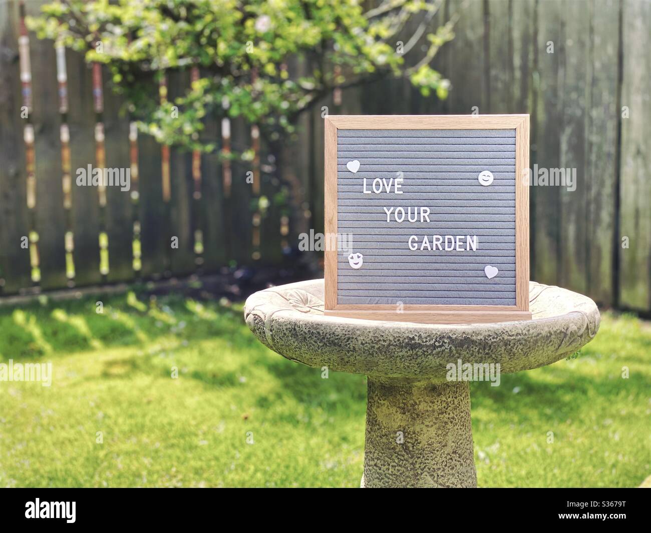 Amate il vostro giardino. Concetto di giardinaggio con bordo di messaggio feltro su un bagno di uccello nel mezzo di un giardino suburbano con l'albero di mela nana nello sfondo e spazio di copia. - Immagine stock catturata con smartphone