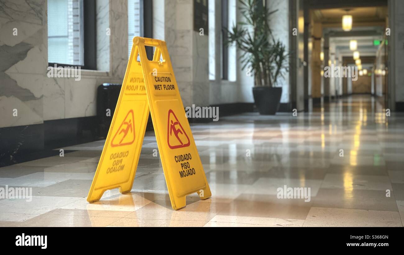 LOS ANGELES, CA, Apr 2020: Cartello giallo 'attenzione, pavimento bagnato' nella lobby piastrellata in marmo con luce naturale proveniente dalle finestre a lato. Corridoio con luci gialle sullo sfondo. Foto Stock