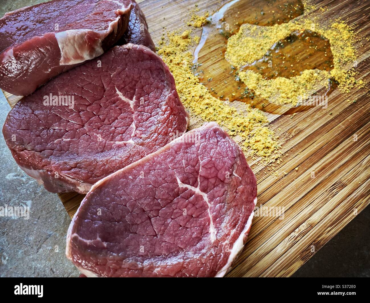 Vista in primo piano delle bistecche di manzo gregge su un rustico tagliere in legno con spezie all'aglio e olio d'oliva. Marinare la carne in cucina prima di cucinare la cena. Foto Stock