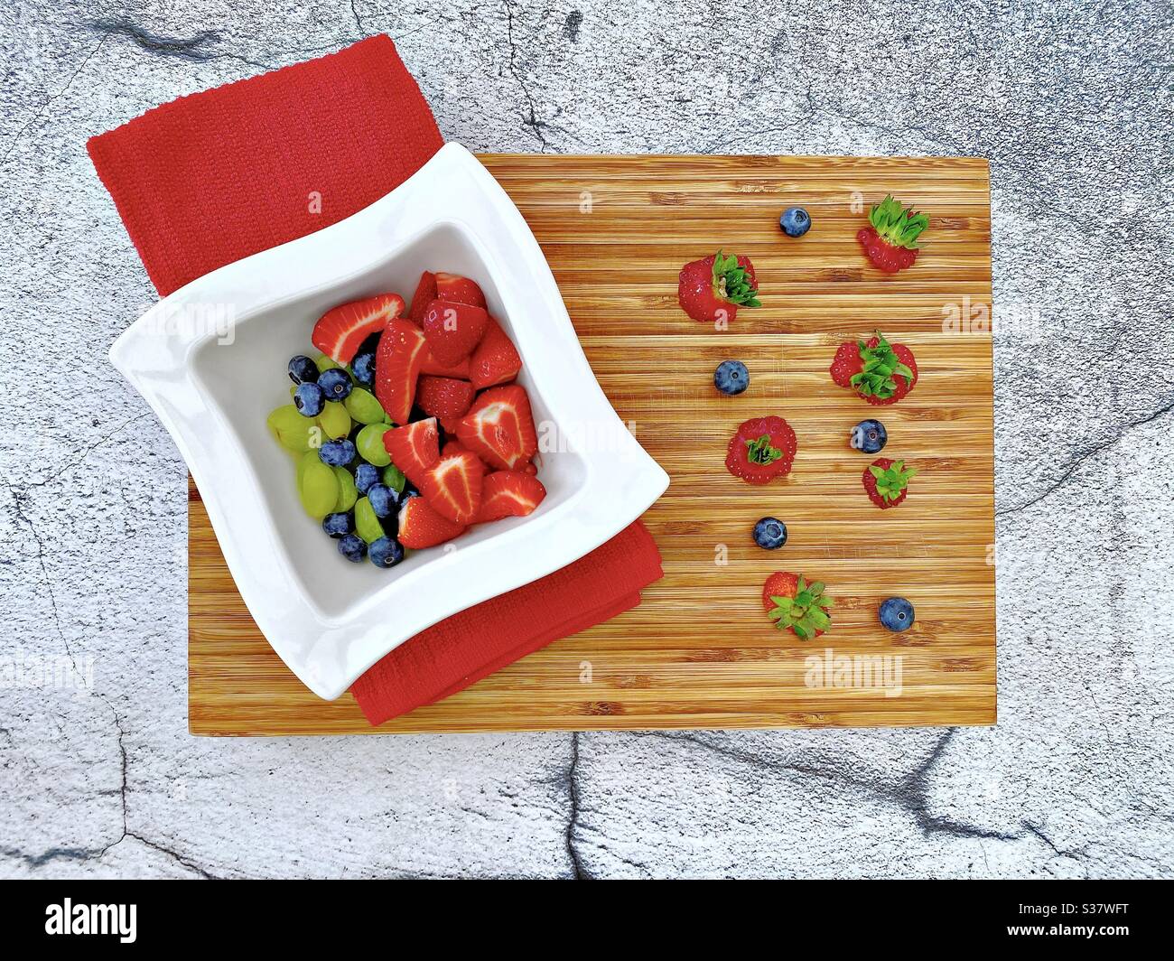 Vista da vicino delle bacche estive fresche in un recipiente bianco contro un rustico tagliere di legno. Preparare fragole, mirtilli e uva sul banco della cucina. - Immagine stock catturata con smartphone