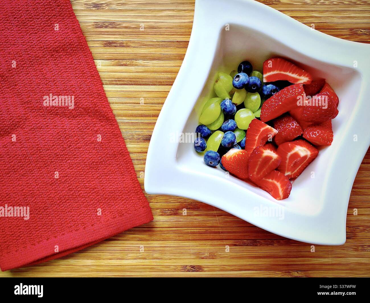 Vista da vicino delle bacche estive fresche in un recipiente bianco contro un rustico tagliere di legno. Preparare fragole, mirtilli e uva sul banco della cucina. - Immagine stock catturata con smartphone