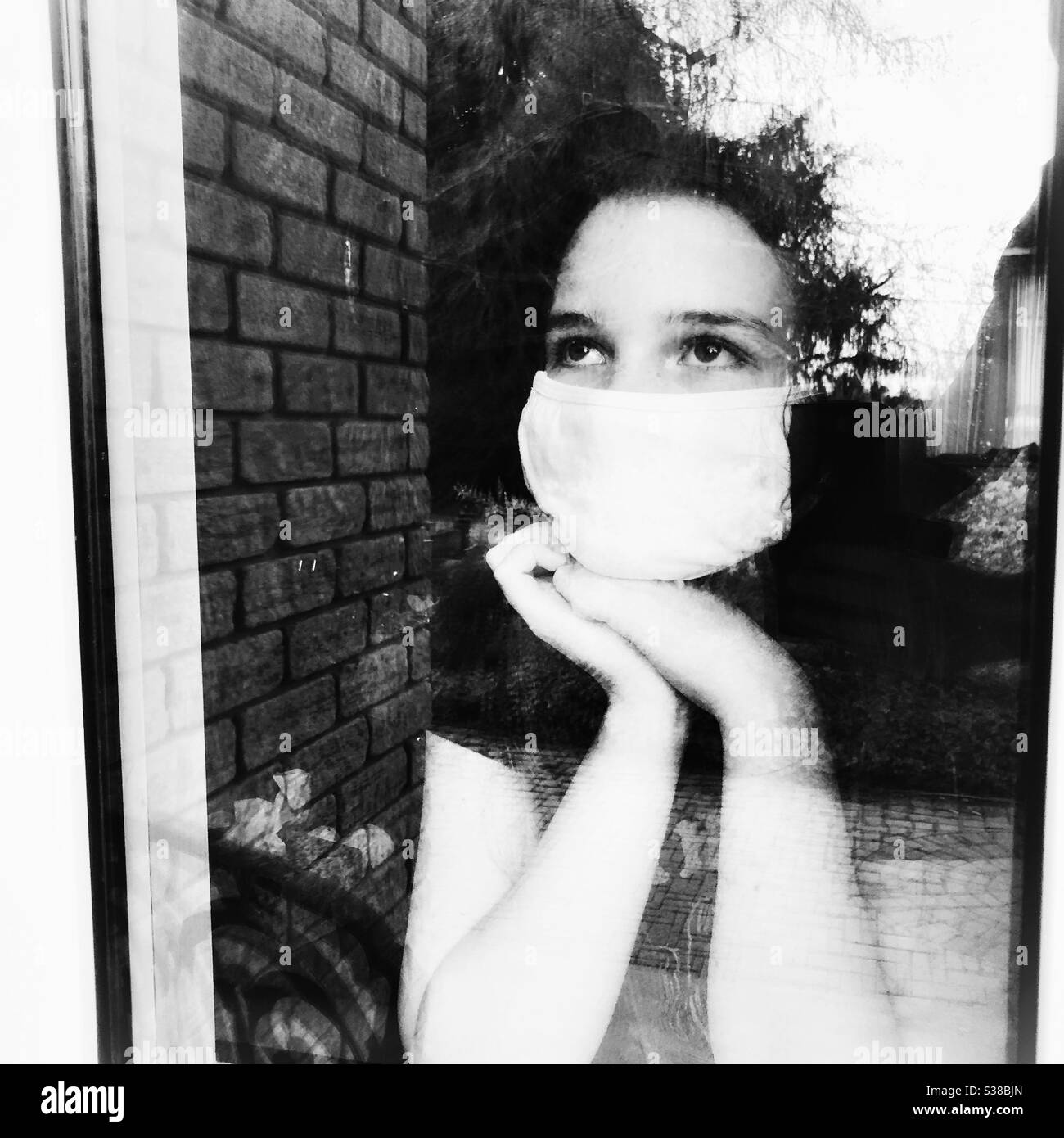 Teen ragazza indossando maschera bianca che guarda fuori la finestra della casa Foto Stock