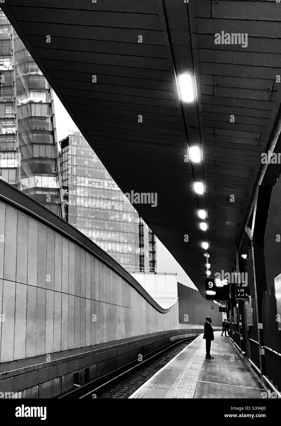 Silhouette di uomo in piedi sulla piattaforma del treno con pista e. edifici dietro Foto Stock